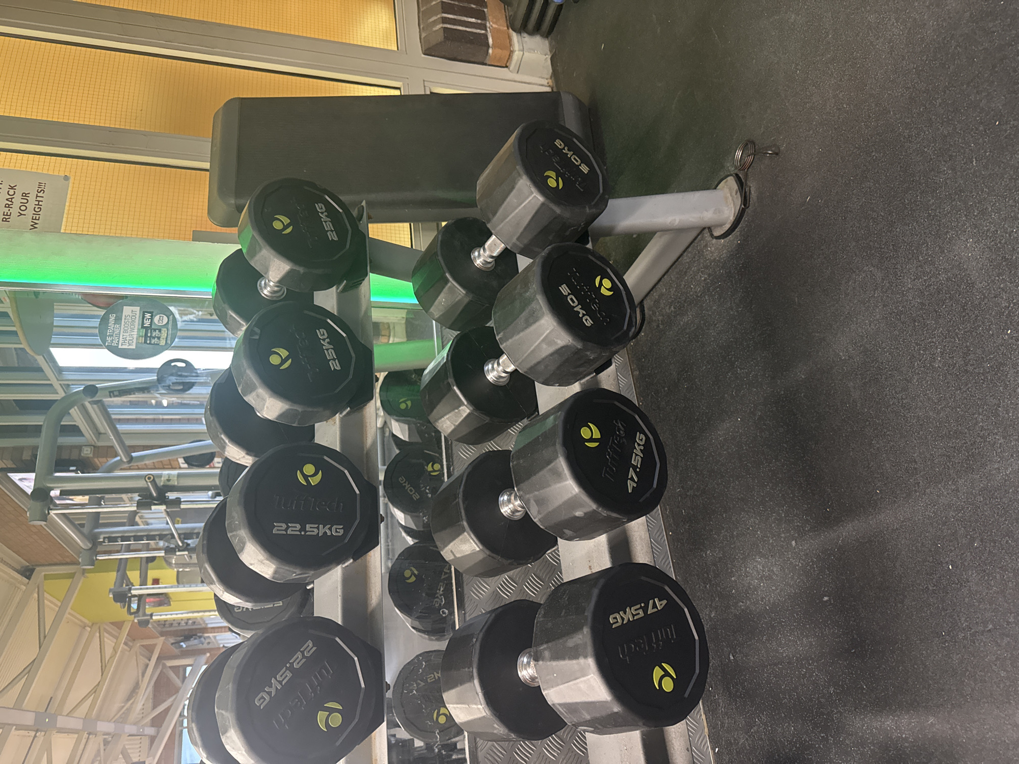 This indoor scene shows a rack of black Tufftech dumbbells, ranging in weight from 22.5KG to 50KG, arranged from light to heavy. The dumbbells are organized on a multi-tiered grey rack. The image includes the text "RE-RACK YOUR WEIGHTS!!!" and "THE TRAINING PARTNER THAT BOOSTS YOUR WORKOUT". The location is a gym in Welshpool, United Kingdom. The environment includes yellow wall decorations.