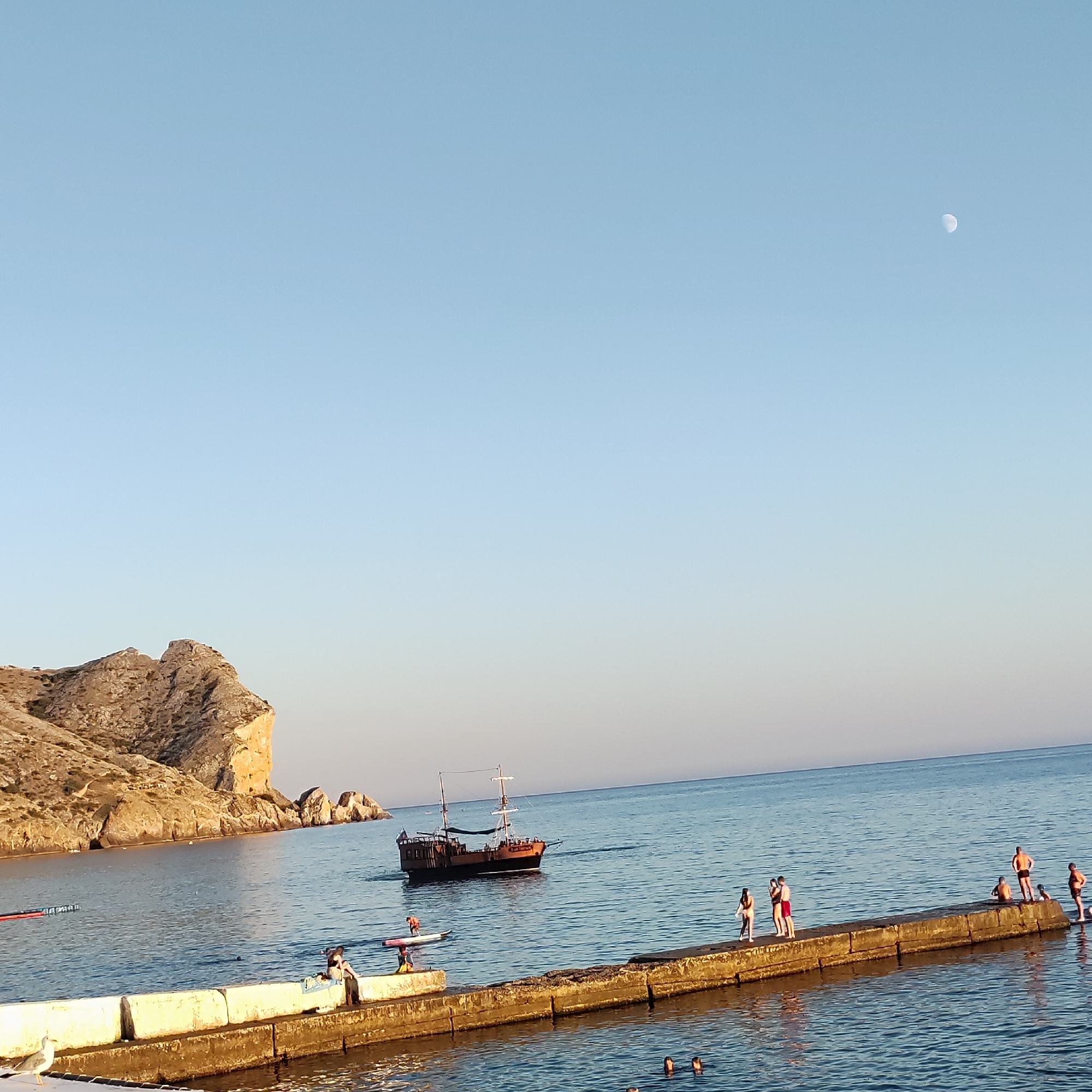 An eye-level, medium shot captures a serene coastal scene under a clear, pale blue sky with a crescent moon visible in the upper right quadrant. The foreground features a weathered concrete pier extending into the calm, blue sea. On the pier, several individuals are present. A woman in a white swimsuit stands with a person in a pink top and white shorts. Further along, a group of men in swim trunks are scattered, some sitting and some standing. Two figures are seen at the edge of the pier, one on a paddleboard wearing an orange vest. In the mid-ground, a wooden sailing ship with two masts is moving away from the viewer towards the left. The backdrop consists of a rugged, tan-colored cliff face on the left side of the frame, with smaller rocky formations in the water near the shore. The water is placid, reflecting the sky and creating gentle ripples. A seagull stands on the concrete edge of the pier in the lower left corner.