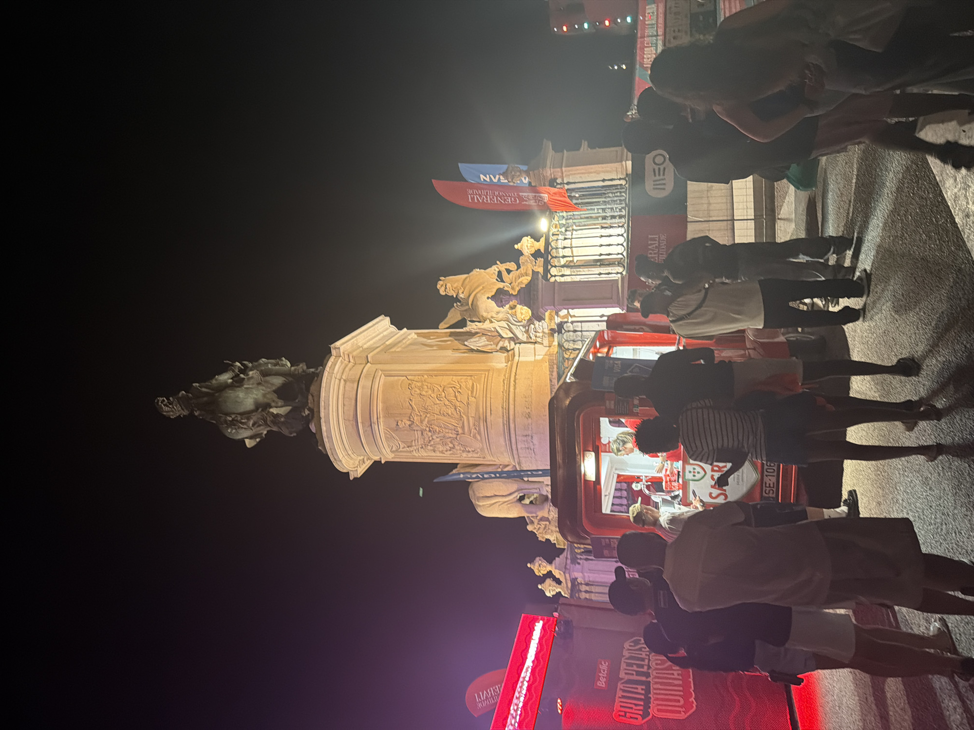 This image shows a night scene in Praça do Comércio, Lisbon, Portugal, featuring a large equestrian statue and a crowd of people. The statue, likely of King José I, stands atop a grand pedestal decorated with bas-reliefs. In the foreground, a red food truck or stall is set up, with people lining up to order. There are several banners and signs visible, including one with "GRITA PELAS QUINAS" and logos for "Betclic" and "VISA". Another banner behind the crowd displays "GENERALI" and "MEO". The ground is paved with characteristic Portuguese cobblestones. The atmosphere appears festive, possibly during an event or celebration.