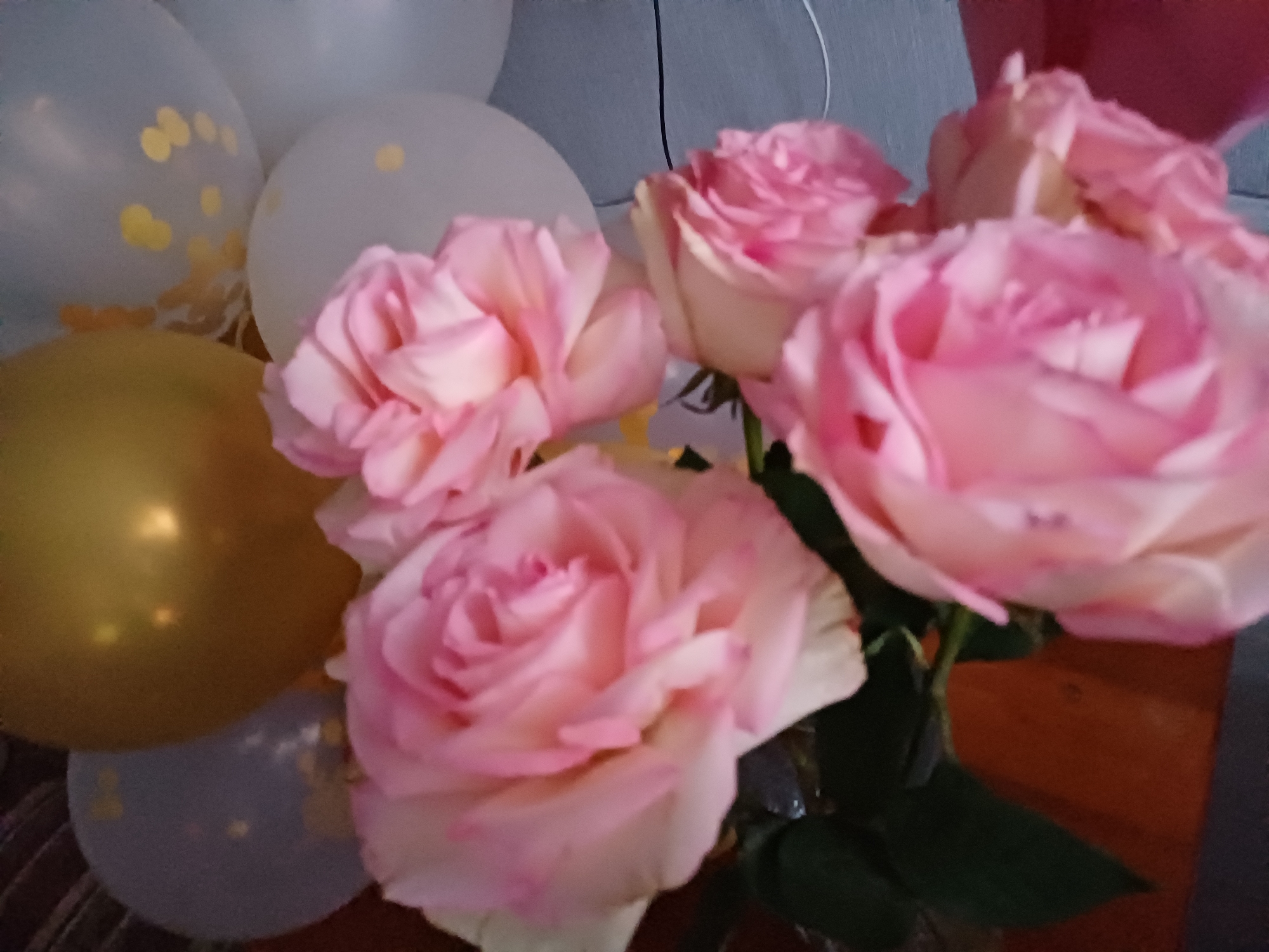 The image depicts a close-up, blurry view of several pink roses and balloons. In the foreground, the soft petals of multiple roses are visible, with varying shades of pink and hints of white. Behind the roses, several balloons can be seen, including one golden balloon and others that appear to be light blue or white with confetti-like patterns. The background is out of focus, showing what appears to be a dark, textured surface. The scene is indoors, and the lighting is soft. There are no people or discernible actions in the image.