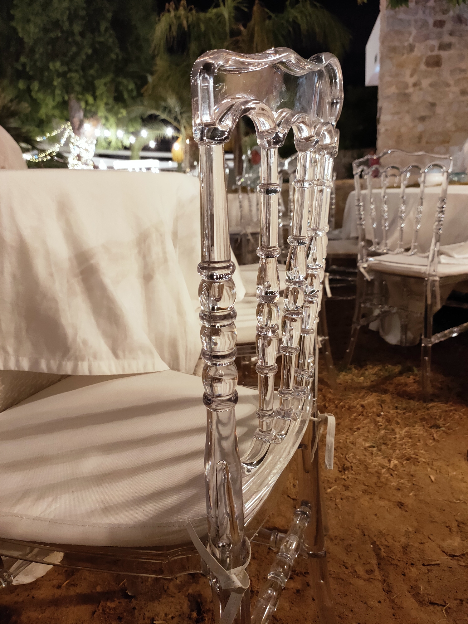 In Cebalat, Tunisia, at night, a series of clear acrylic chairs are arranged around a table covered with a white cloth. The chairs have a decorative design, with segmented legs and a curved backrest. The ground beneath the chairs appears to be dirt and straw, suggesting an outdoor setting. In the background, trees are visible, illuminated by string lights, and a stone building is partially visible. The scene appears to be set up for an event, possibly a wedding or reception. There are no people in the immediate foreground, but indistinct figures can be seen in the background.