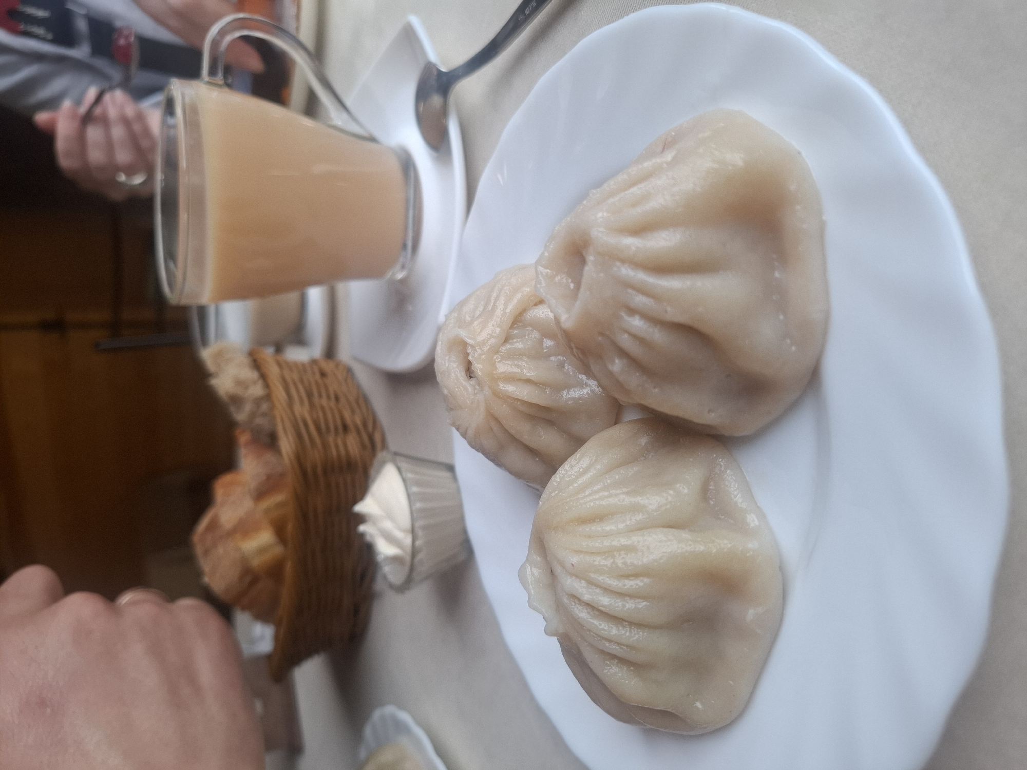 A close-up, eye-level shot shows a white plate with three steamed dumplings arranged in a triangular pattern. The dumplings are pale and have a pleated top. To the left of the plate is a small glass dish containing a dollop of white sauce. Behind the plate, a woven basket contains sliced bread. Further back, a clear glass mug filled with a light brown beverage, likely tea with milk, sits on a saucer. A spoon rests on the saucer. In the background, out of focus, a person's hand is visible, and another person's hands are holding a spoon with a dollop of red food. The scene appears to be set in a cafe or restaurant, possibly in Sukhoy Ruchey, Russia, given the context.