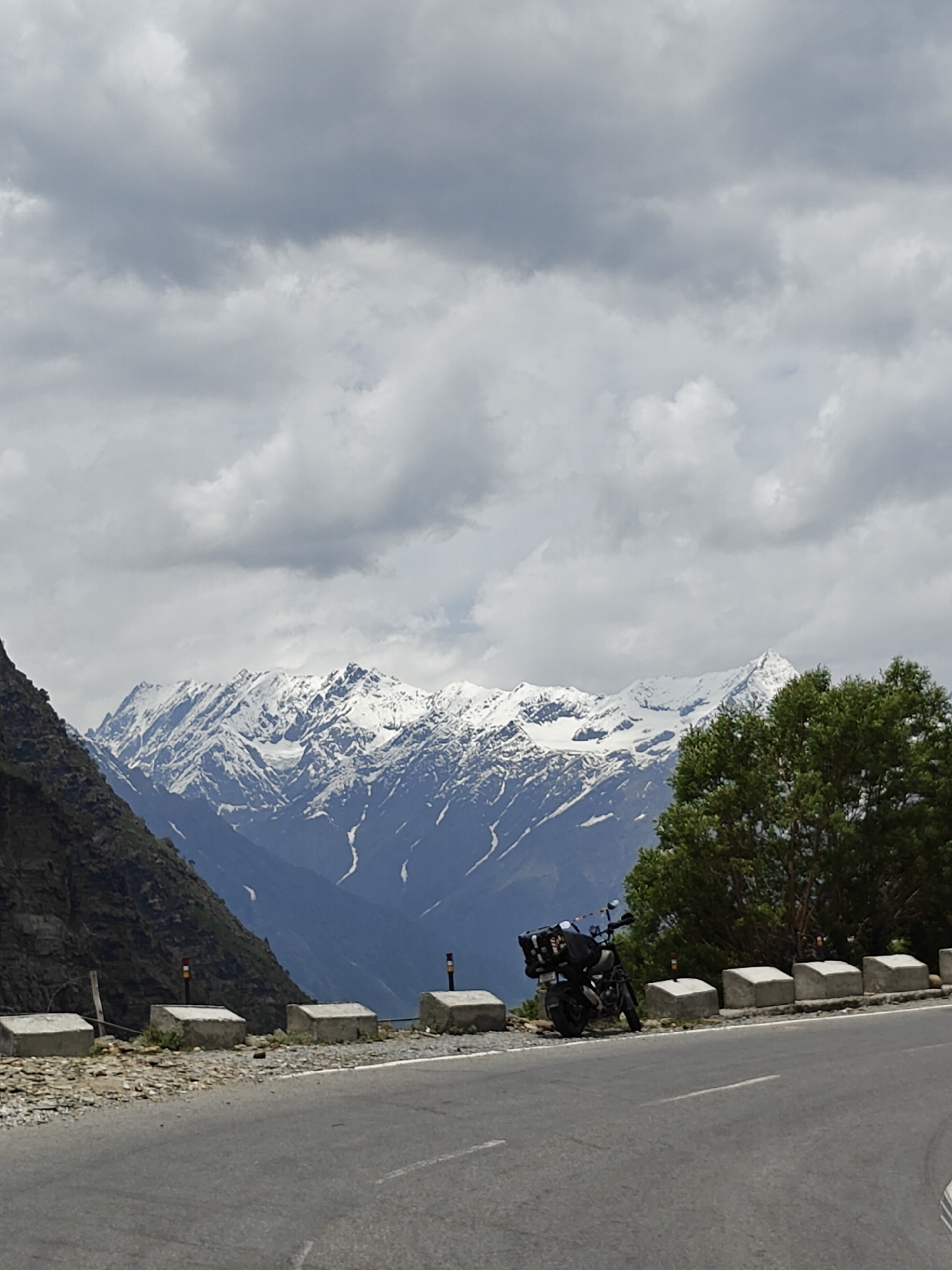 A motorcycle is parked on the side of a paved mountain road in an area with snow-capped mountains in the background. The motorcycle, a black touring model, is loaded with luggage. The road has a white line marking the center. Concrete barriers line the edge of the road, some with reflective markers. A steep, rocky incline is visible on the left side of the road. Lush green trees are on the right side of the road, obscuring some of the view of the mountains. The sky is filled with heavy gray clouds.