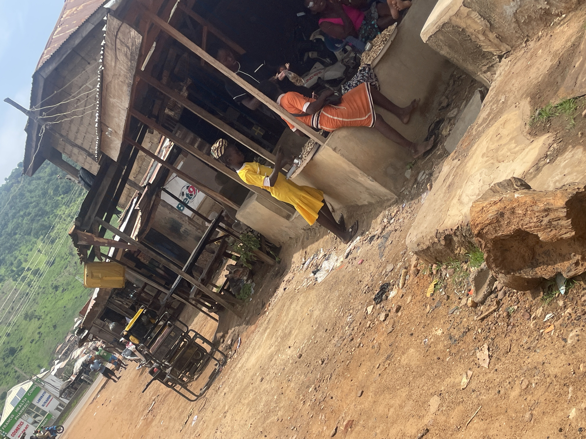 The image depicts an outdoor market scene in Lokoja, Nigeria. In the foreground, a dirt path littered with debris leads to a makeshift wooden structure with a rusted corrugated metal roof, serving as market stalls. Several people are visible within and around these stalls. Two young girls are at the forefront of the stalls, one wearing a bright yellow dress and the other an orange dress. They appear to be interacting with goods displayed on raised platforms. Behind them, other individuals, including adults, are also present, some appearing to be vendors or shoppers. To the left of the stalls, a motorcycle and a generator are parked on the dirt road. In the background, a building with the signage "ST. FRANCIS HEALTH CONSULTANCY PHARMACY" is visible, and beyond that, green, hilly terrain characteristic of Lokoja. The overall scene suggests a bustling, informal market environment in a semi-urban setting.