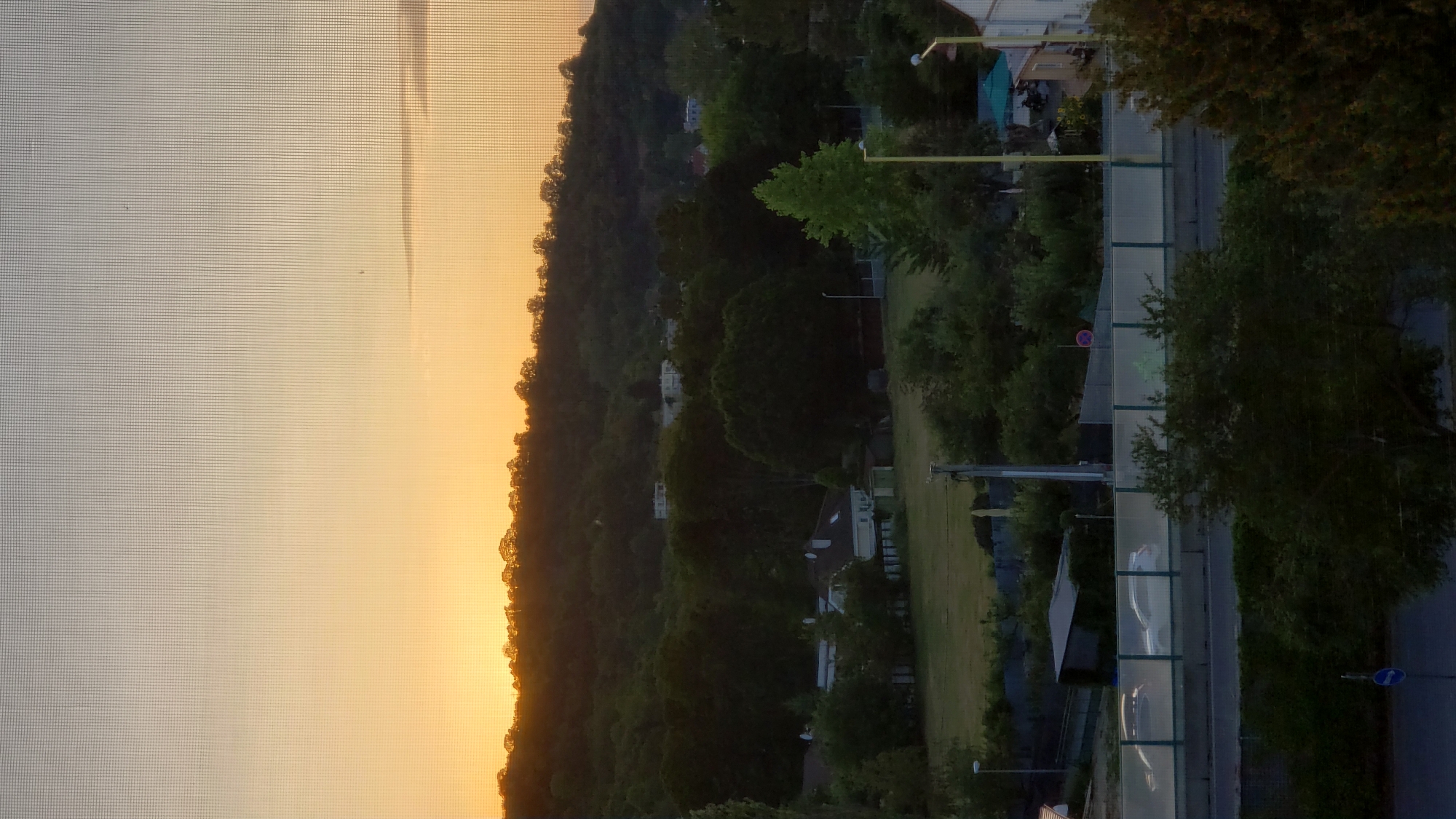 The image captures a sunset over a residential area in Prešov, Slovakia, viewed through a window screen. The sky transitions from a bright orange hue near the horizon to a pale grey at the top. A silhouetted tree line crowns a gently sloping hill in the mid-ground, with scattered buildings visible amongst the trees. Below the hill, a field is bordered by dense foliage and houses. A road with a sound barrier is in the foreground, with two cars driving on it. The overall scene depicts a tranquil evening in a suburban environment.