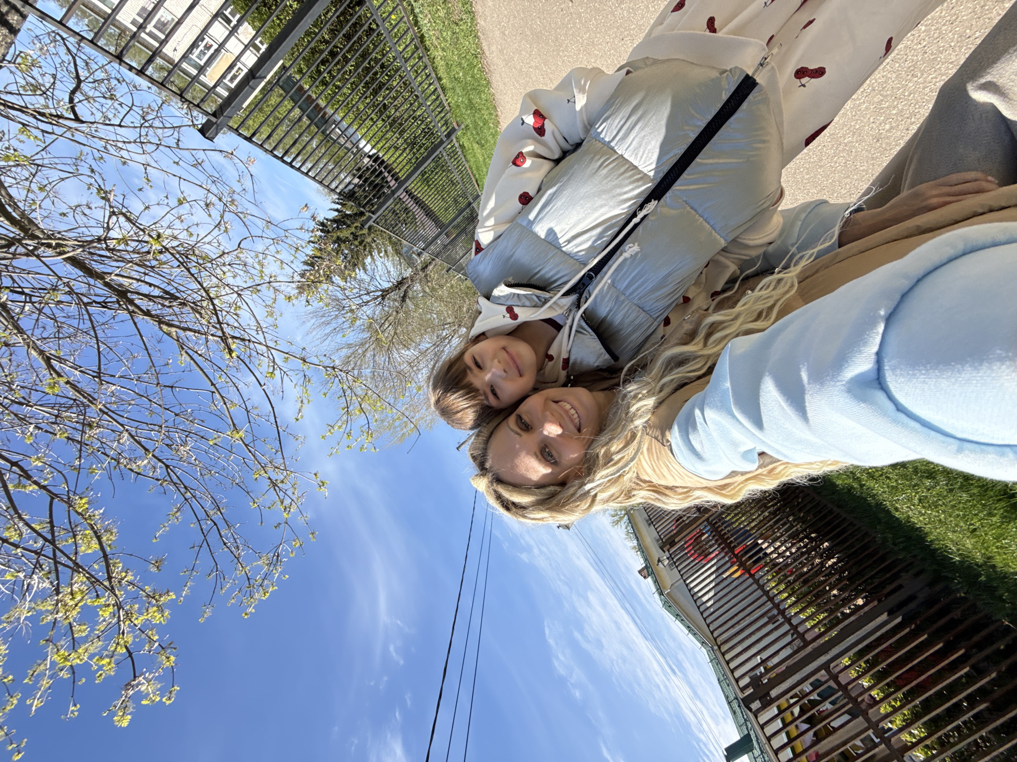 A woman and child are posing closely for a selfie on an urban street. The woman is likely blonde. The child is wearing a white hooded jacket with a cherry pattern, and a shiny puffy jacket. The background includes a blue sky with light clouds, bare tree branches, utility lines, fencing, grass, and building elements. While the exact location in Заволжье, Russia cannot be determined from the visual information, it is evident they are in a city.