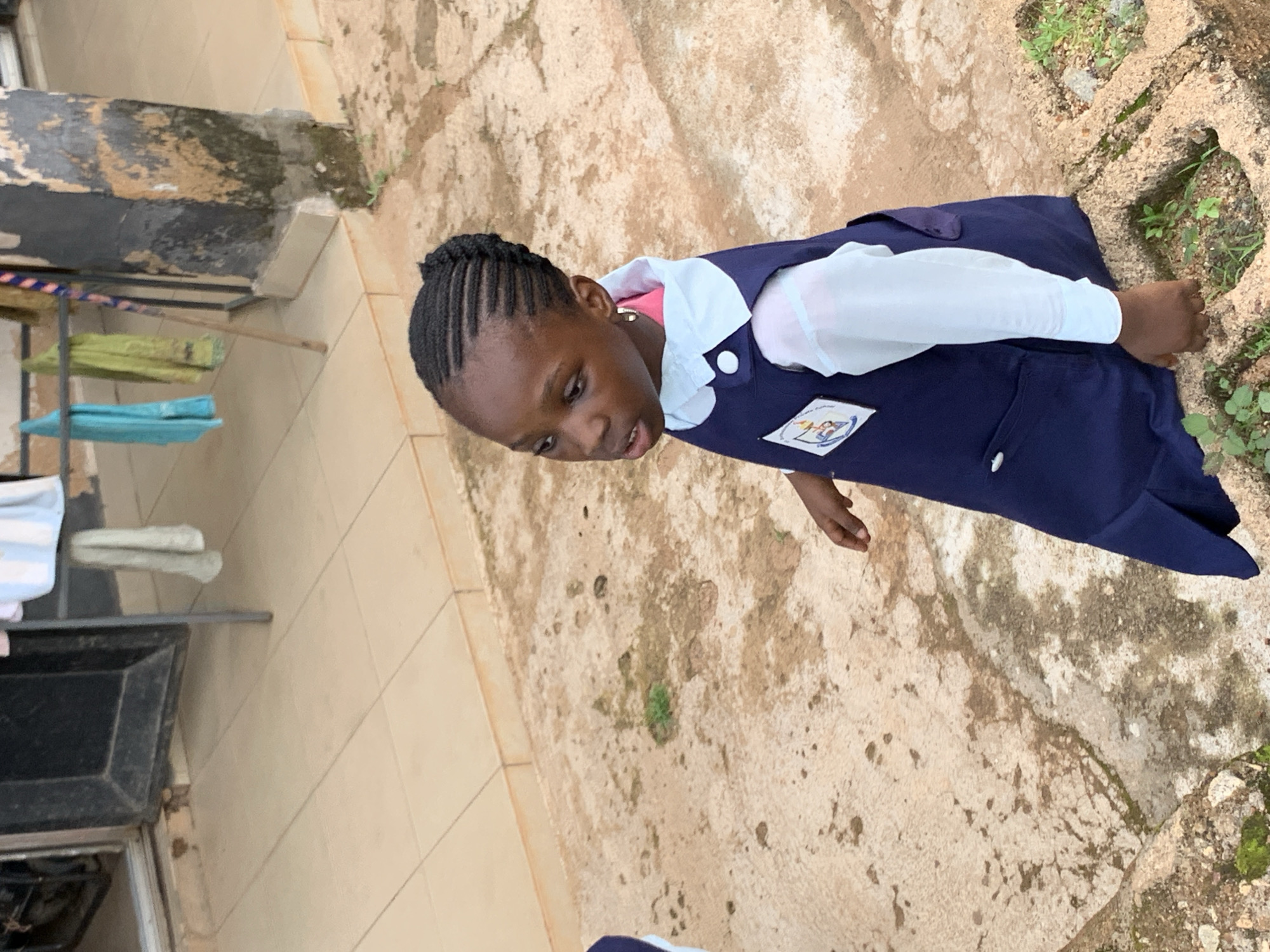 A young Black girl, likely a student, is captured in a medium-full shot, her gaze directed downwards and to the left. She wears a white long-sleeved shirt beneath a dark blue pinafore dress, complete with two white buttons at the shoulder. A patch, likely a school emblem, is visible on the left side of the pinafore. Her hair is styled in neat braids.
The setting appears to be an outdoor courtyard with a rough, mottled ground surface, possibly concrete or packed earth, interspersed with small patches of green vegetation. To the left of the frame, a weathered, dark-colored structure, possibly a wall or building facade, shows signs of decay with peeling paint or plaster. Adjacent to this structure is a rack holding several items of clothing, including what appears to be a light blue cloth and a white garment.
The lighting suggests it is daytime, with diffused light, indicating either overcast conditions or a shaded area. The overall mood is observational, focusing on the child in her school attire within a seemingly ordinary outdoor environment. The location context of Jos, Nigeria, places the scene within a specific geographical and cultural setting. No legible text is visible in the image.