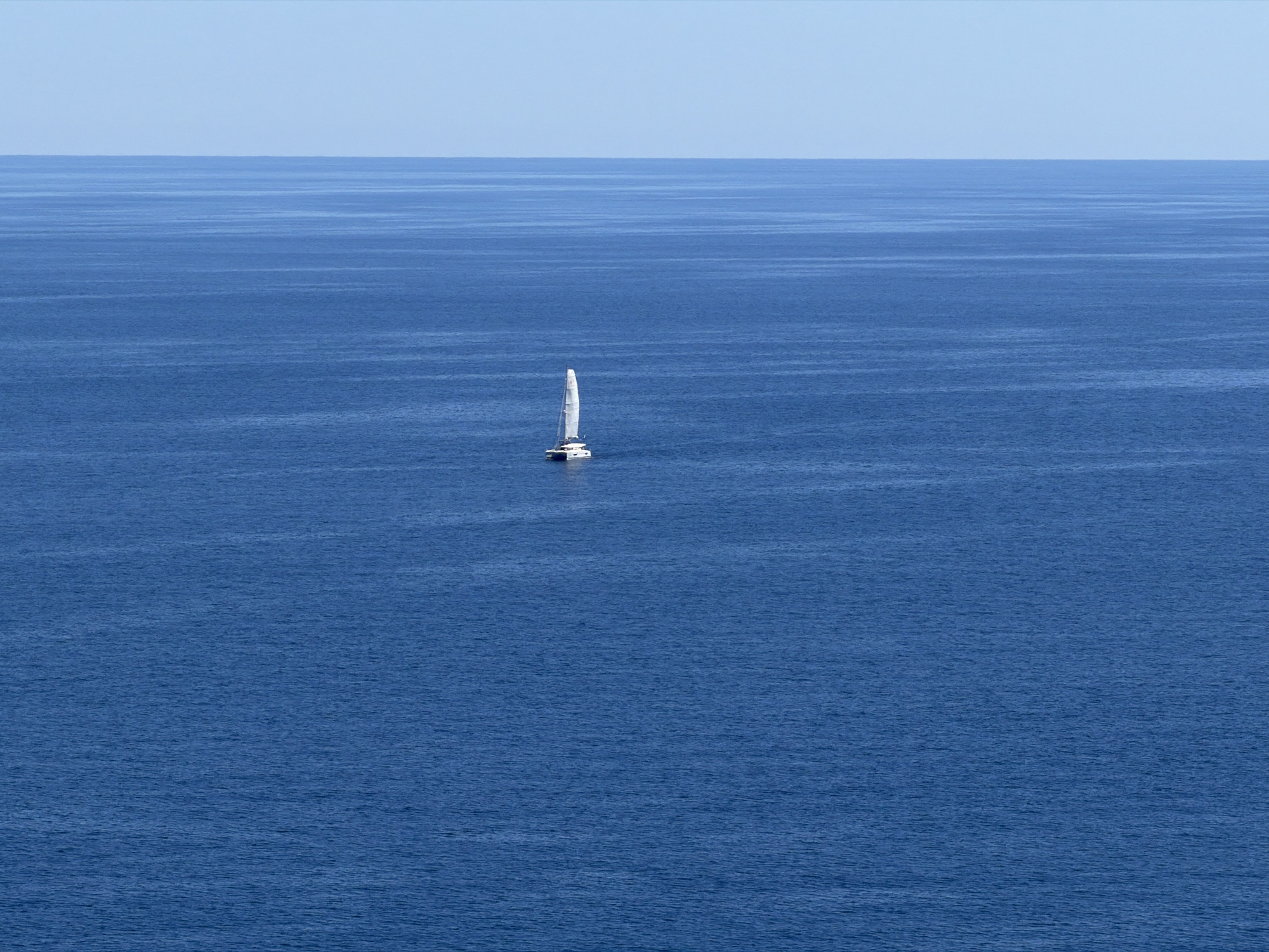 The scene is a daytime ocean view in Capdepera, Spain. A catamaran sailboat with a white sail is located near the center of the image. The ocean is deep blue with subtle wave patterns. The sky is a pale blue, meeting the ocean at the horizon line in the background.