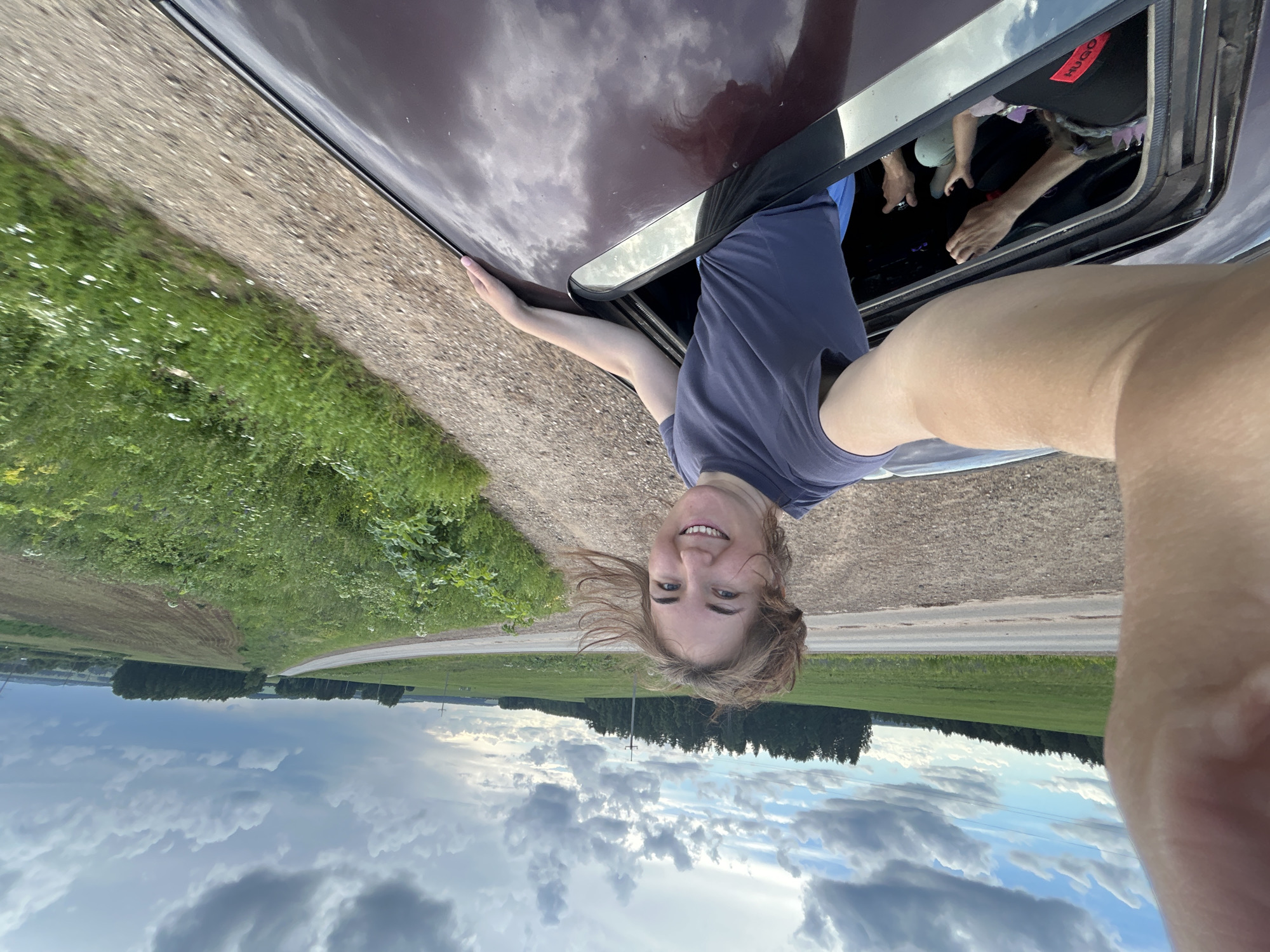 The image captures a young woman leaning out of a car window, with her upper body and head visible. She is smiling and appears to be enjoying herself. The camera is held from her perspective, creating a selfie-style shot. Her left arm is extended outside the car, resting on the vehicle's roof. Inside the car, other individuals are partially visible.
The background shows a rural landscape with green fields and a cloudy sky, suggesting an outdoor setting during the daytime. The car appears to be on a gravel road or a dirt track next to a grassy area. The reflection of the sky and clouds on the car's dark, glossy surface is noticeable.
The image is oriented sideways, with the subjects appearing upside down. The clothing of the woman is a plain dark t-shirt. A red tag with the word "HUGO" is visible on a black item of clothing inside the car. The overall mood seems lighthearted and adventurous, possibly indicating a road trip or a scenic drive. The location context provided is Верх-Рождество, Russia.
