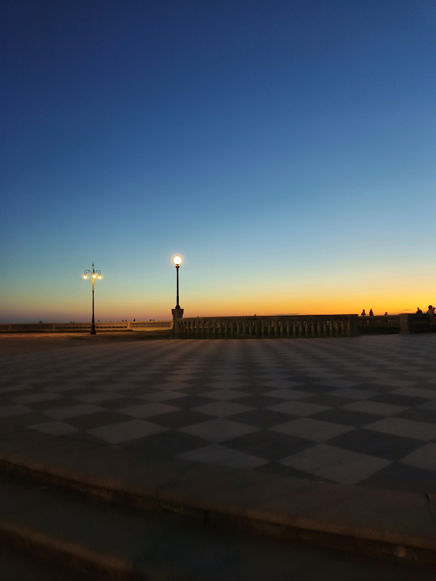 The image depicts the Terrazza Mascagni in Livorno, Italy, at dusk. A checkered pavement dominates the foreground, with diamond-shaped tiles in varying shades of grey. A stone balustrade, featuring decorative elements, stretches across the middle ground. Two ornate lampposts are visible: one on the left with three lights and another on the right with a single, illuminated bulb. Silhouettes of several people can be seen along the balustrade, some appearing to stand and observe the sunset. The sky transitions from a deep blue at the top to a vibrant orange and yellow horizon, indicating the setting sun. The overall atmosphere is serene, capturing the tranquil beauty of the iconic seaside promenade as daylight fades.
