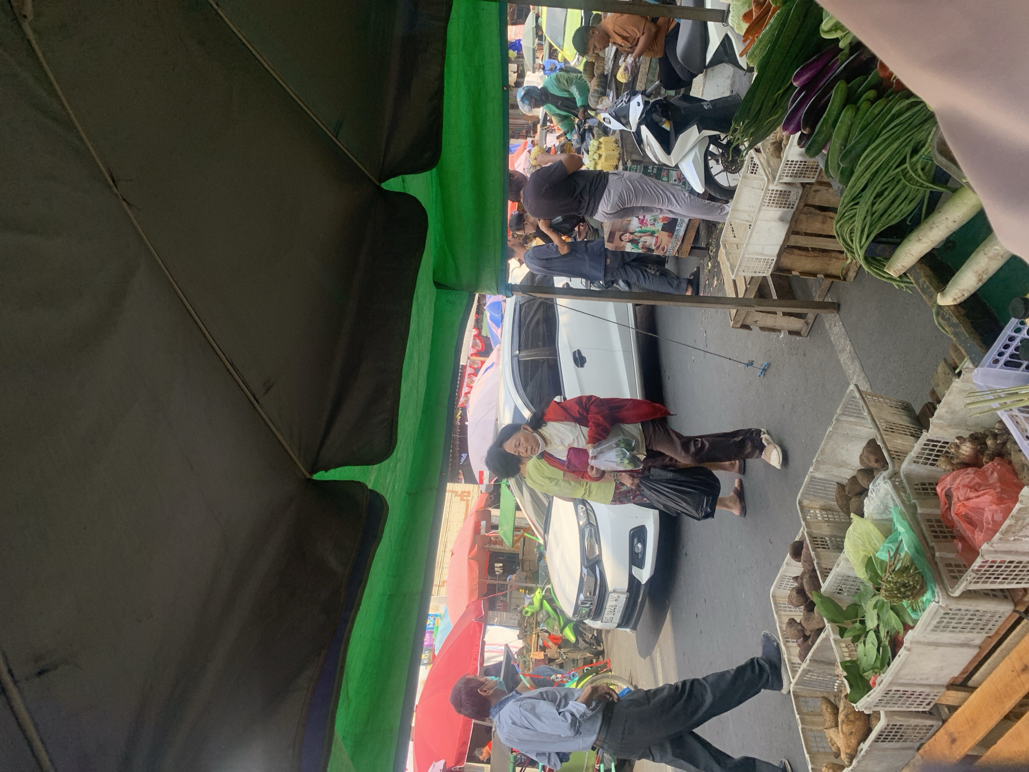 This image depicts a bustling street market scene in Palembang, Indonesia, likely near a road. In the foreground, several white plastic crates are overflowing with fresh produce, including long beans, cucumbers, eggplants, radishes, and what appear to be ginger roots and taro roots. A wooden pallet is visible below some of the crates, and a partially visible arm and shoulder in the lower right corner suggest the photographer is also at the market.
Several people are present and engaged in various activities. A man wearing a light blue shirt and dark pants, with a blue face mask, walks towards the left of the frame. In the middle ground, two women are walking; one in a red jacket and brown pants carries a plastic bag of produce, while the other, in a green shirt and patterned skirt, walks beside her. A white car is parked behind them. Further back, several motorbikes and stalls with colorful umbrellas (red and green) are visible, indicating a vibrant marketplace. People are seen interacting with vendors, browsing produce, and riding motorbikes. The overall environment suggests a typical outdoor market on a bright, possibly sunny day, with a mix of pedestrian and vehicle traffic.