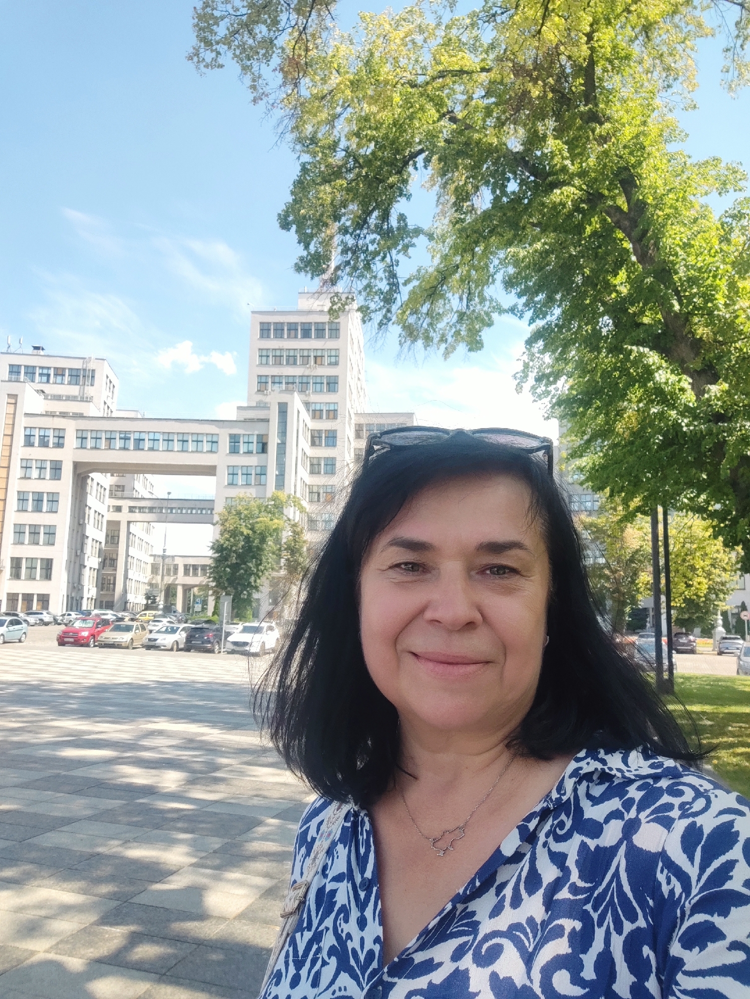 In this medium shot selfie taken outdoors, a woman with shoulder-length black hair and dark eyes smiles directly at the camera. She is wearing a blue and white patterned blouse and a delicate silver necklace. Black sunglasses are perched on top of her head. Behind her, a large, modern white building with many windows is visible, connected by an elevated walkway. The building appears to be part of a city square or plaza, with a paved ground and several parked cars in the background. Trees with green foliage are present on the right side of the frame, and the sky is blue with scattered clouds. The lighting suggests it is a sunny day.