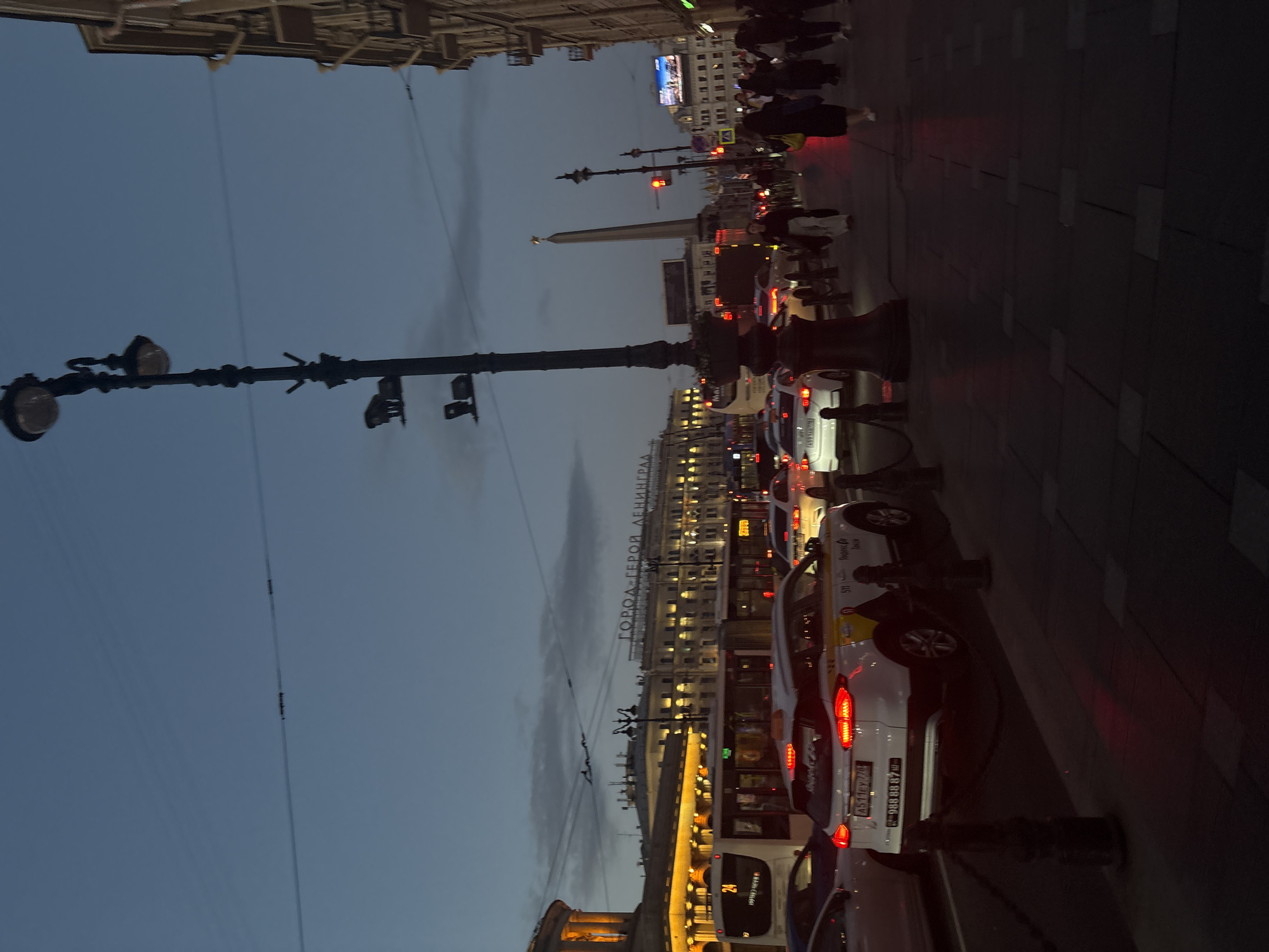 The image depicts a street scene in Saint Petersburg, Russia, likely at dusk or early evening, given the dim lighting and illuminated buildings and streetlights. The view is from a sidewalk looking across a busy street.
Dominating the left side of the frame is a tall, ornate street lamp with multiple lights. Beyond the street, several vehicles are visible, including a white bus with the number "24" displayed on its front, and a white SUV with yellow and black markings, appearing to be a taxi. Other cars are present in traffic, with their brake lights illuminated. The street is lined with buildings. A prominent building in the background features illuminated windows and a large sign that reads "ГОРОД-ГЕРОЙ ЛЕНИНГРАД" (City-Hero Leningrad). To the right of this building, a tall obelisk-like monument with a star on top is visible.
On the sidewalk in the foreground, several pedestrians are walking. They are mostly silhouetted against the dim light. The sidewalk is paved with stone tiles. The overall atmosphere is that of a well-lit urban environment experiencing evening traffic.
