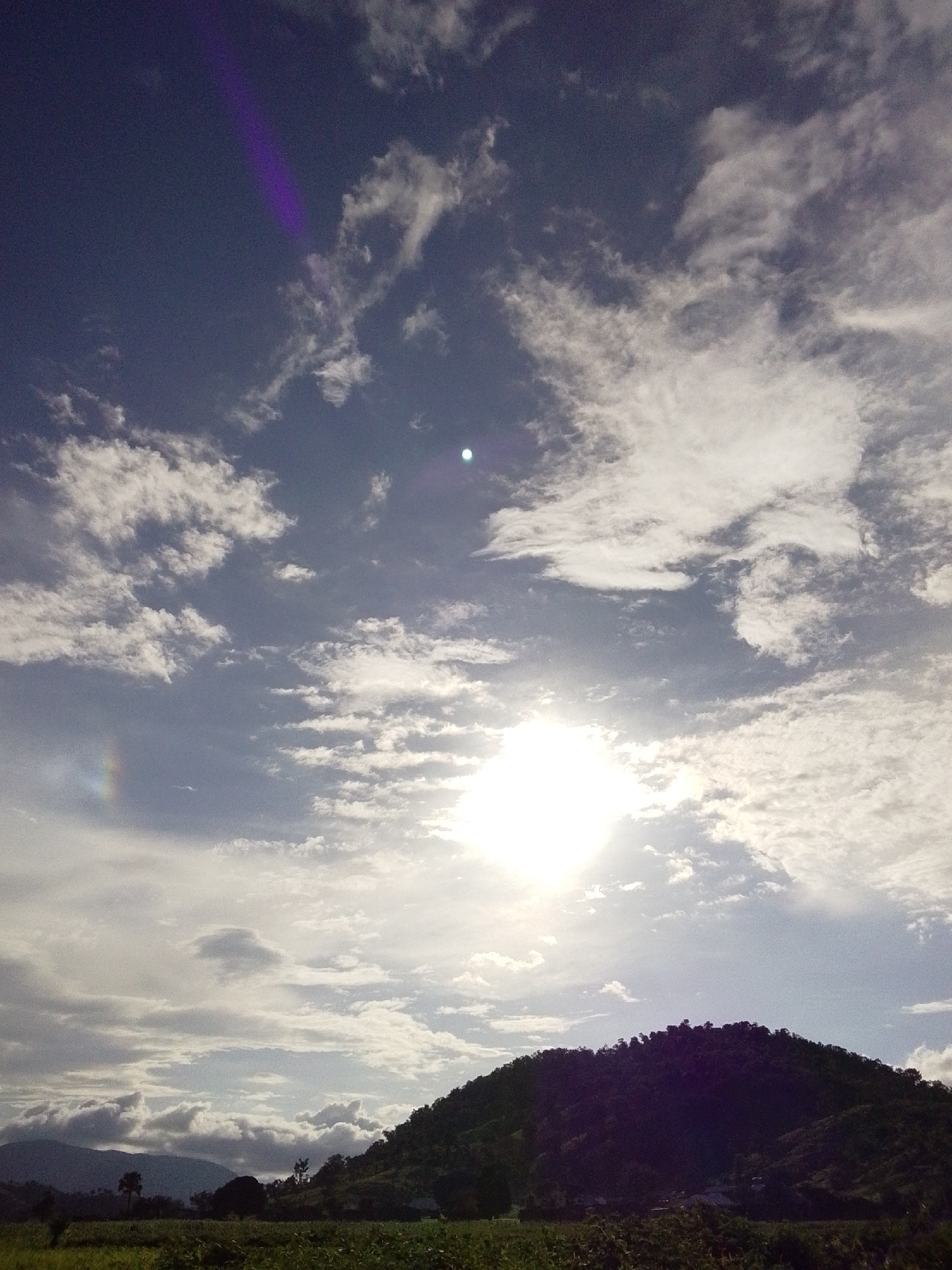 This is a photograph of a sunlit sky over a hilly landscape in Serti, Nigeria. The sky is filled with scattered white clouds, with the sun appearing as a bright, blown-out orb near the center of the frame. A faint purple streak is visible in the upper left portion of the sky, possibly a lens flare or atmospheric phenomenon. To the left of the sun, a subtle rainbow arc can be seen.
The lower half of the image shows a dark, densely vegetated hillside that dominates the right side. The base of the hill is bordered by a field of green crops or vegetation, and further back, there are silhouetted trees and distant, hazy mountains on the left. The overall lighting suggests it is daytime, likely late afternoon or early morning given the angle of the sun and the dramatic contrast between the illuminated sky and the shadowed landscape. The scene conveys a sense of natural beauty and the vastness of the sky. There is no visible text in the image.