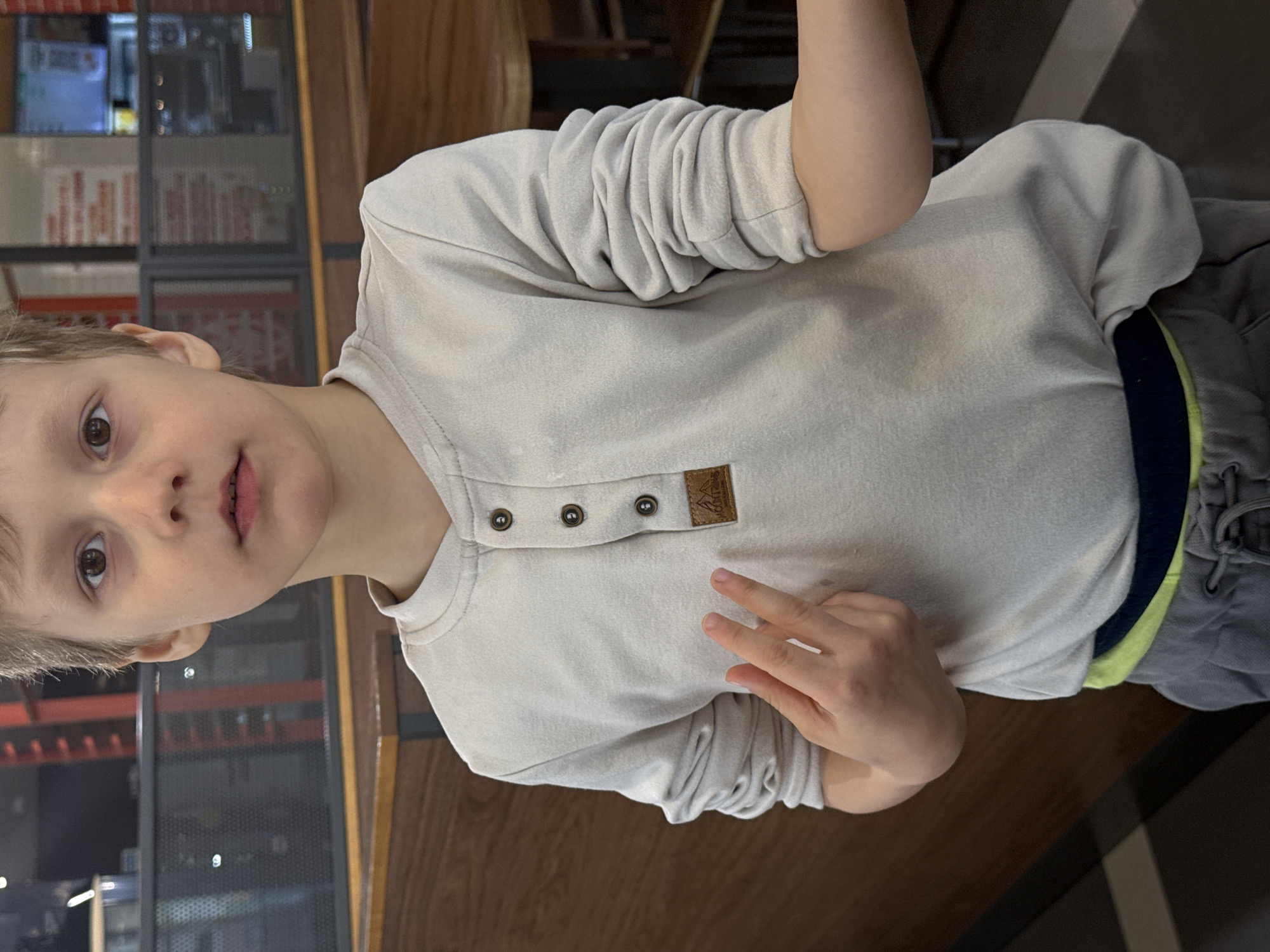 An image, rotated 90 degrees counter-clockwise, depicts a young boy indoors in Tobolsk, Russia. The boy, who appears to be around early primary school age, has light-colored hair and expressive brown eyes. He is looking directly at the viewer with a neutral to slightly curious expression, his lips slightly parted.
He is wearing a light-beige or off-white long-sleeved shirt with a Henley-style placket featuring three dark brown buttons. On the right side of his chest, there is a small, rectangular brown patch, possibly leather or faux leather, with an embossed design that appears to include a mountain shape and some faint, illegible text. His sleeves are pushed up, creating visible folds. He is also wearing grey sweatpants with a distinct neon green and dark blue stripe visible at the waistline. His left hand is raised, showing two fingers (a peace sign or indicating the number two).
The background suggests an indoor public setting, possibly a casual restaurant, cafe, or store. Visible elements include dark wooden surfaces, which could be furniture or wall panels, and what appears to be glass partitions or shelving units with dark metal frames. Through these structures, various blurred objects and light reflections are discernible. The lighting is artificial, common for indoor spaces. There is no clearly readable text besides the ambiguous patch on his shirt.