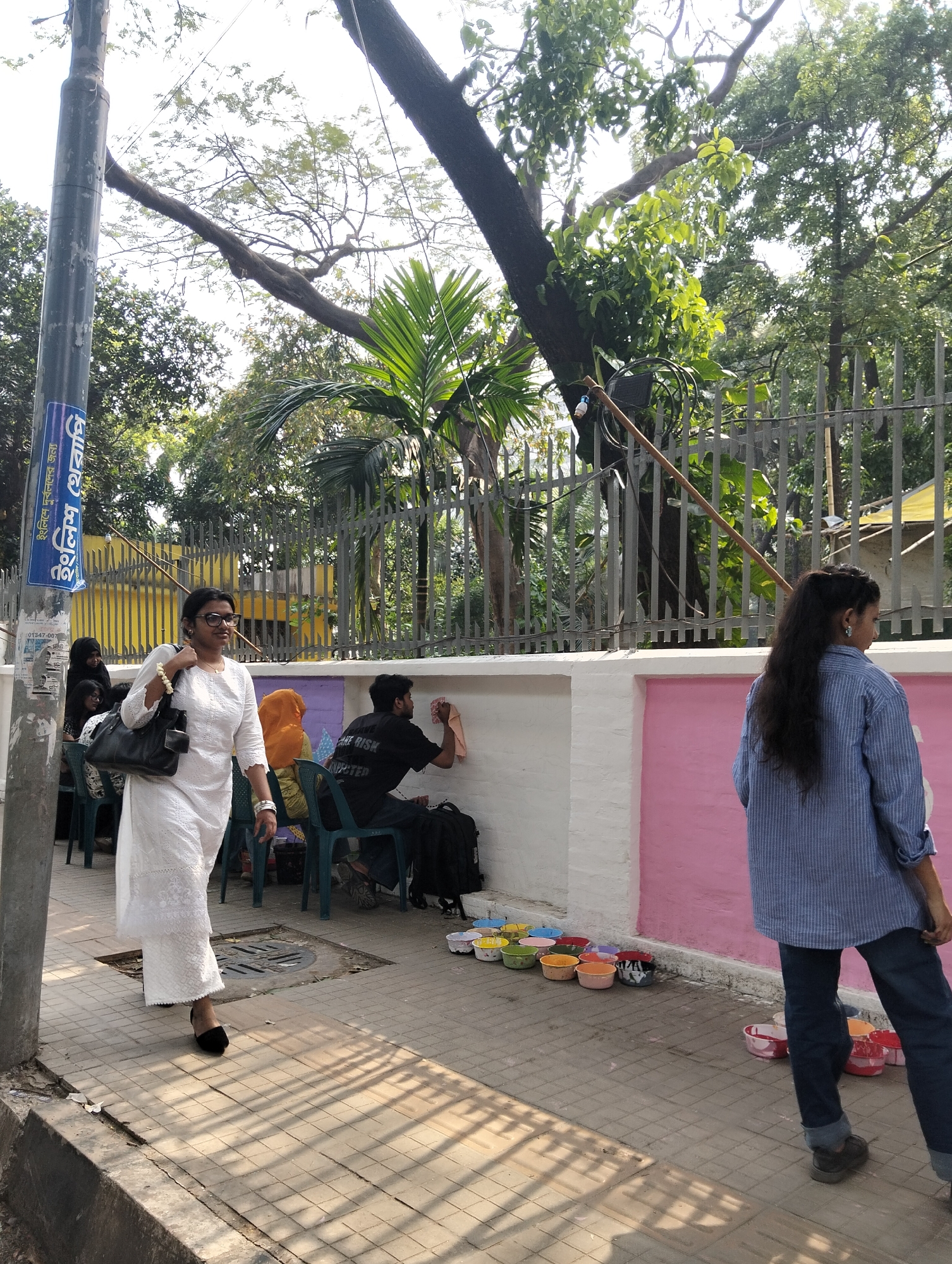 A street scene in Dhaka, Bangladesh. A woman in a white dress and black shoes walks along the sidewalk. Another woman in a blue striped shirt and jeans walks in the opposite direction. A row of people sit in chairs, some wearing hijabs. A man in a black t-shirt cleans graffiti off a white and pink wall. Small colorful bowls line the base of the wall. A metal fence separates the sidewalk from a park or yard with trees and vegetation. A utility pole stands on the left side of the image.