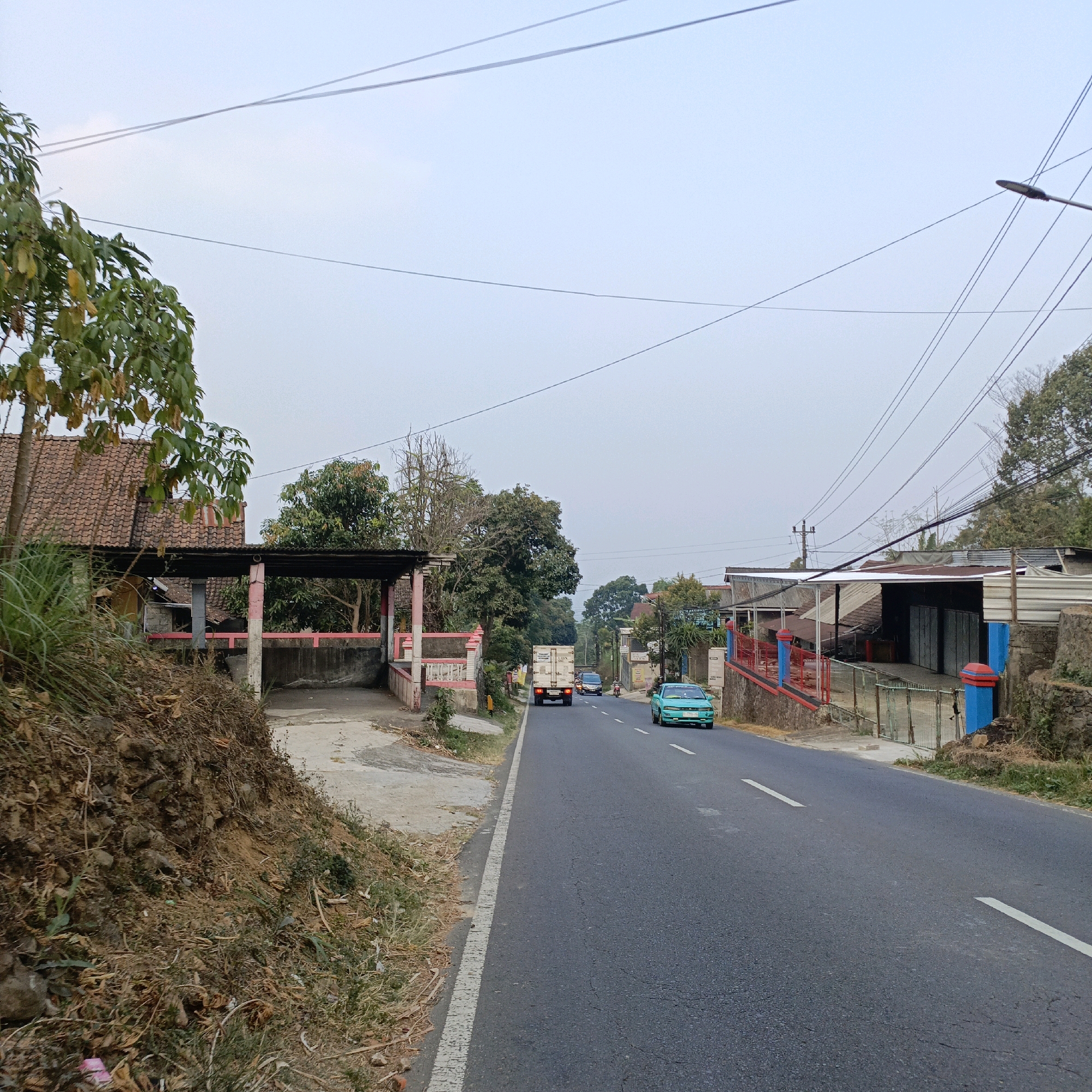 The image depicts a rural road scene in Sumogawe, Indonesia. A two-lane asphalt road runs from the foreground into the distance, curving slightly to the right. The sky is overcast and a light blue. Overhead, numerous power lines crisscross the frame.
On the left side of the road, there is a steep, dirt embankment covered in dry grass and some sparse vegetation. Adjacent to the embankment, there is a building with a red tile roof and a covered carport area supported by columns.
On the right side of the road, there are several buildings with corrugated metal roofs, some of which appear to be shops or small businesses. A red railing with blue posts runs along the front of one of these buildings.
Several vehicles are present on the road. A white box truck is traveling away from the camera in the center lane. Ahead of the truck, two cars are visible, one dark-colored and one a light green sedan, also traveling away from the camera. A motorcycle is also seen further ahead, in the right lane. The surrounding vegetation consists of trees and various plants. The overall impression is that of a typical roadside environment in a semi-rural Indonesian setting.