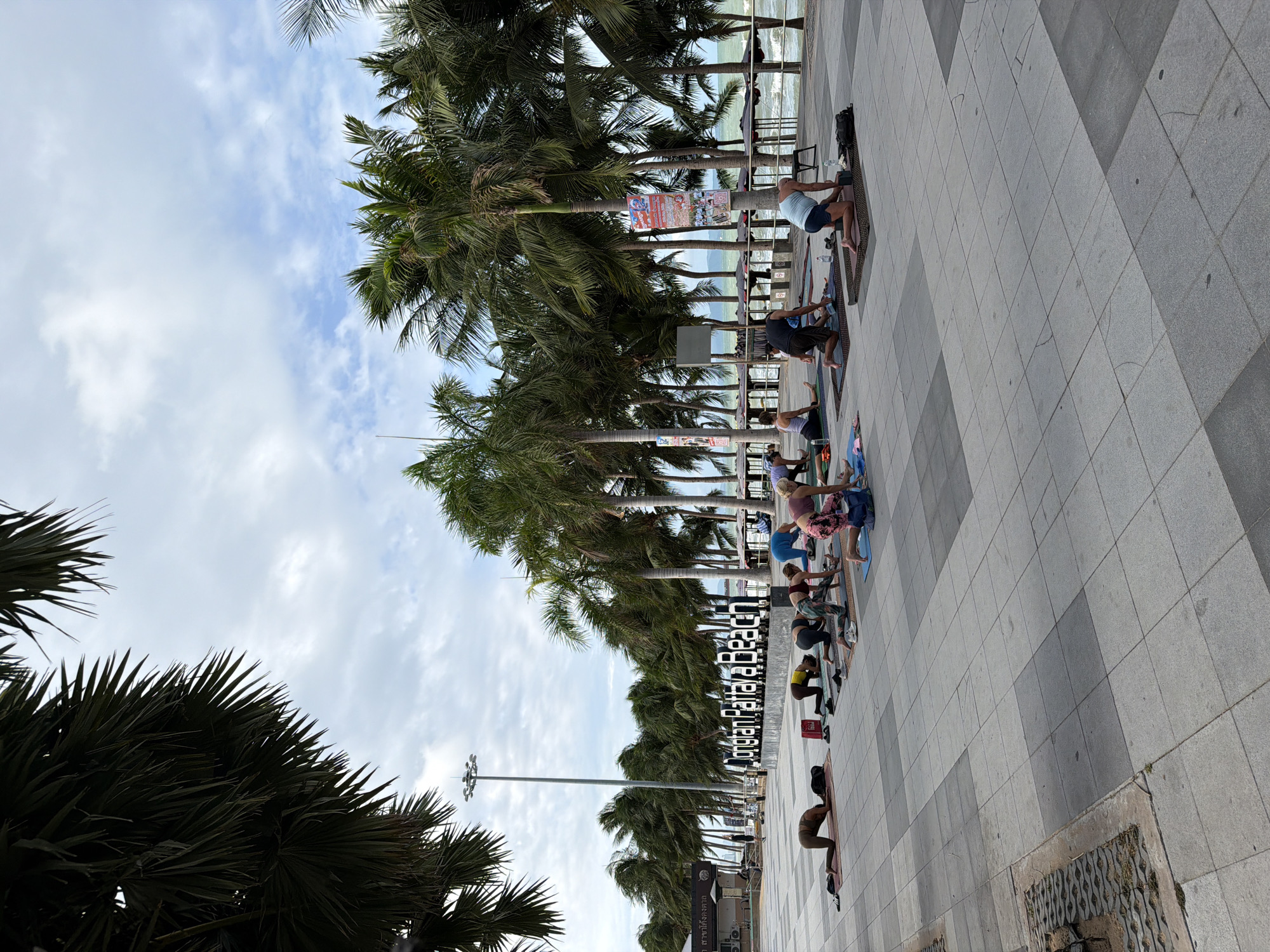 This image depicts an outdoor yoga session on the paved promenade of Jomtien Pattaya Beach in Pattaya City, Thailand. Approximately seven to nine individuals, predominantly adults in athletic wear, are observed performing yoga poses on individual mats laid out on the light and dark grey tiled surface. Most participants are in a kneeling or quadruped position. Personal items such as water bottles, shoes, and small bags are visible near their mats.
In the mid-ground, a prominent sign with large, dark letters reads "Jomtien Pattaya Beach." A long line of tall palm trees extends across the background, bordering the promenade and the beach. A tall, grey light pole with multiple lamps is situated behind the sign. To the right, beyond a barrier or railing, the sea with visible waves can be seen. The sky is partly cloudy, displaying both blue patches and white clouds. Additional structures and a colorful poster affixed to a palm tree are also visible in the scene.