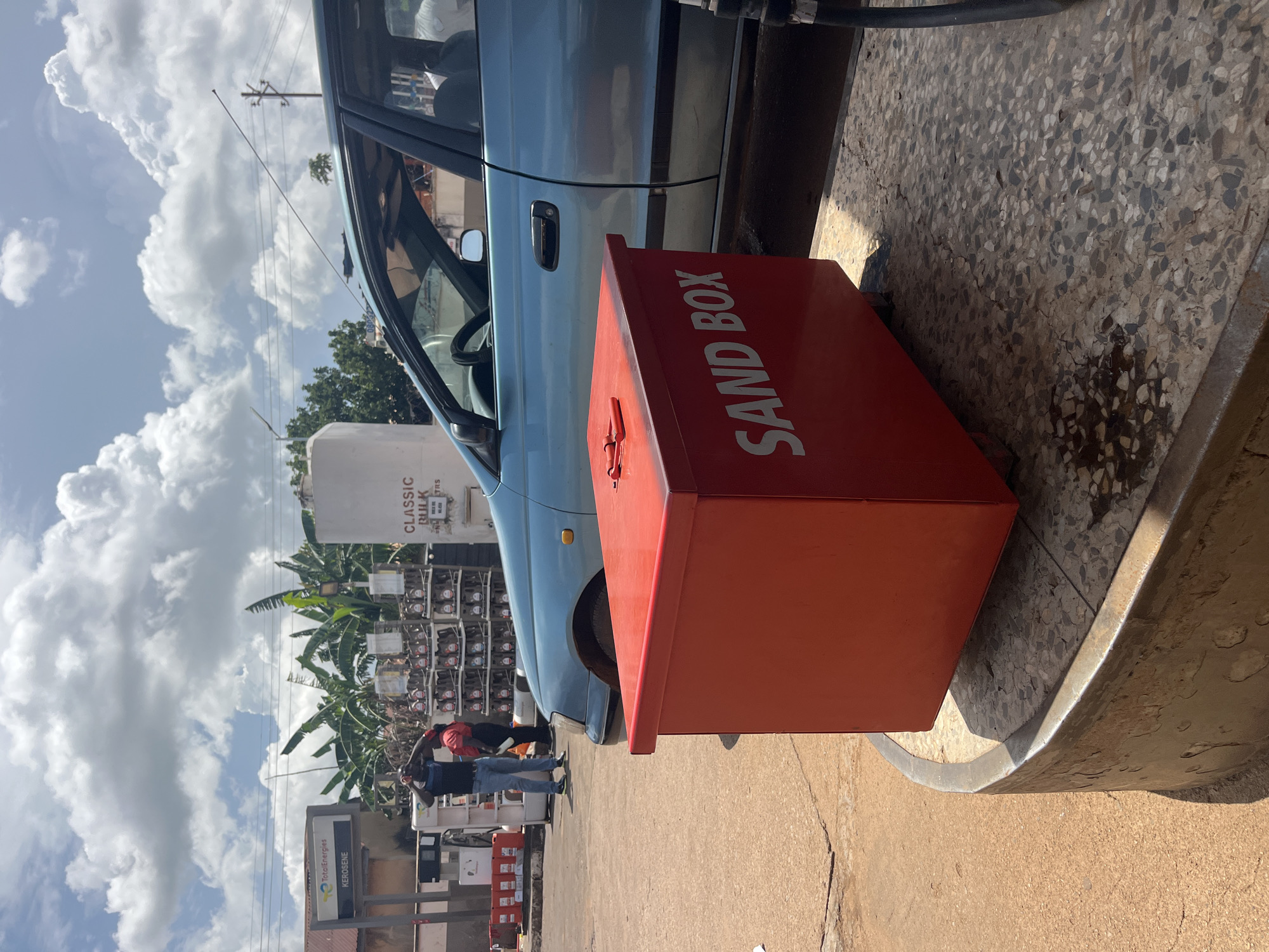 In Gura Top, Nigeria, a bright red sandbox is positioned on a textured concrete platform in front of a light blue car. The sandbox has the words "SAND BOX" printed in white on its side. Behind the sandbox, a light blue car is parked, with its driver's side door open. In the background, a gas station is visible, with a sign that reads "TotalEnergies KEROSENE." Several people are present at the gas station, some appearing to be attendants and customers. The sky is cloudy, with patches of blue visible. There are also several racks of oil bottles and a large white tank in the background. The overall scene suggests a typical day at a service station in a Nigerian city.