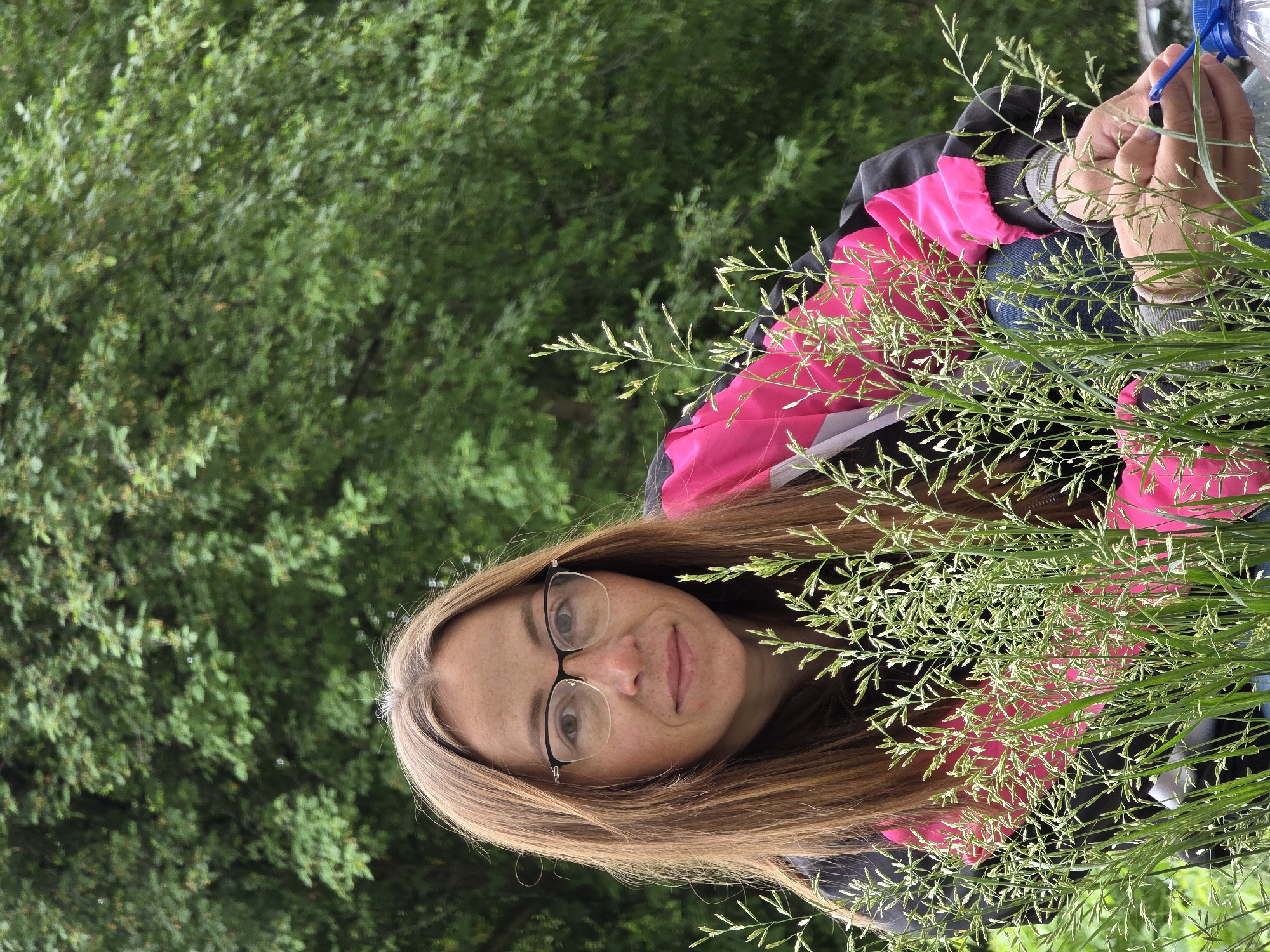 A woman with light brown hair and glasses is pictured in a field of tall grass. She is wearing a dark jacket with pink accents and blue jeans. Her hands are visible, holding a blue plastic object near a plastic water bottle. The background consists of green trees and foliage. The location is identified as Chaikovsky, Russia.
