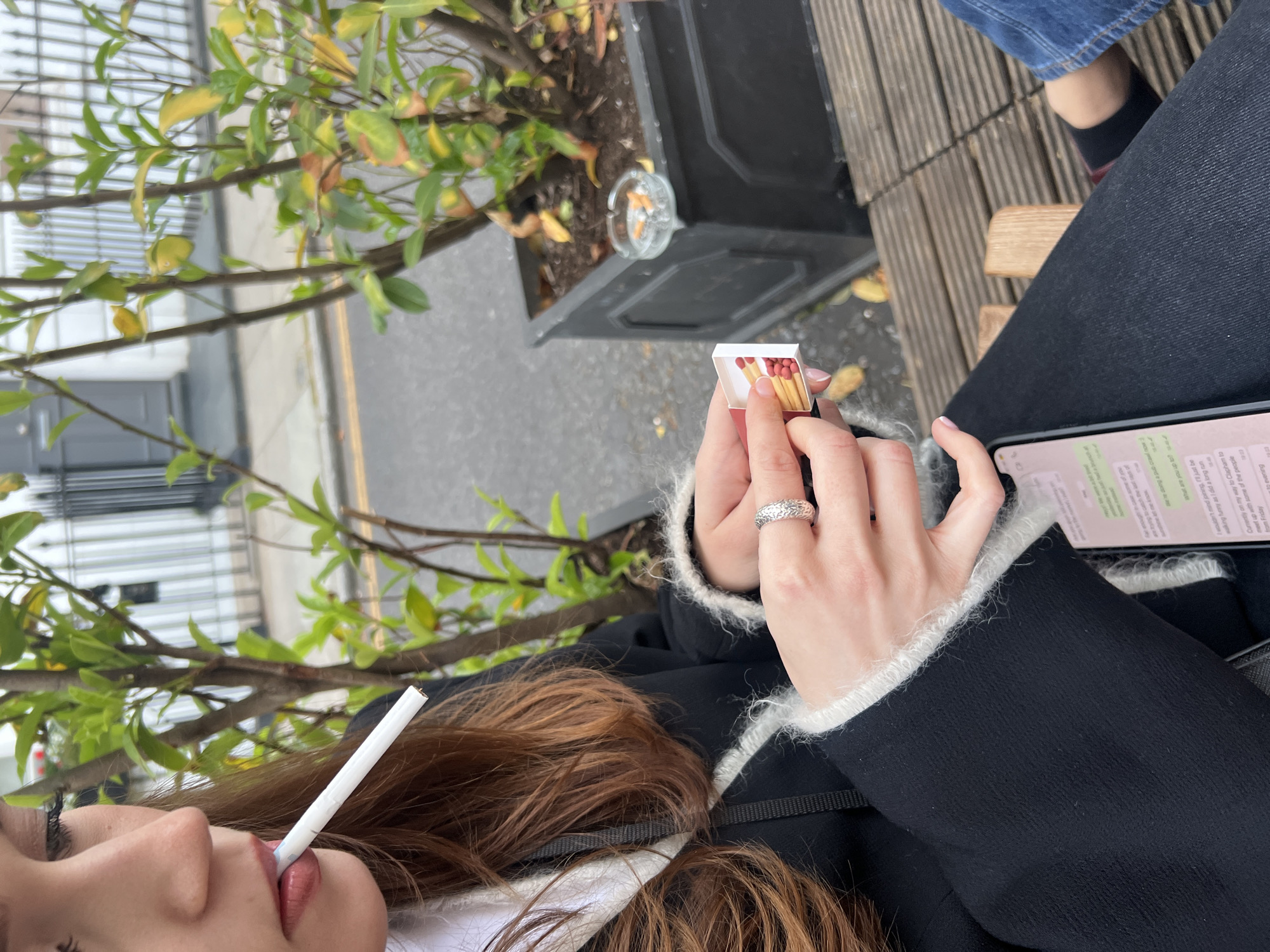 A person is sitting outdoors, holding a lit cigarette in their mouth and a box of matches in their hand. They also hold a smartphone with a WhatsApp conversation visible on the screen. An ashtray filled with cigarette butts is located near a plant in a pot. The location appears to be urban, possibly London, as indicated by the WhatsApp message referring to "Clapham" (a district in South London).