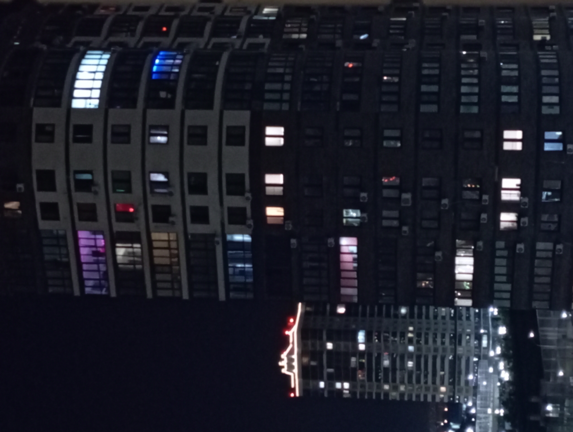 The image shows a night view of two high-rise apartment buildings in an urban environment, possibly in the city of Unknown, Unknown. The building on the right is taller and has a curved facade, while the building on the left is rectangular and has decorative lighting on its roof. Numerous windows on both buildings are illuminated with various colors, including white, pink, and blue, suggesting occupancy. The sky is dark and clear. The scene depicts a typical urban landscape at night, with artificial lighting from residential buildings.