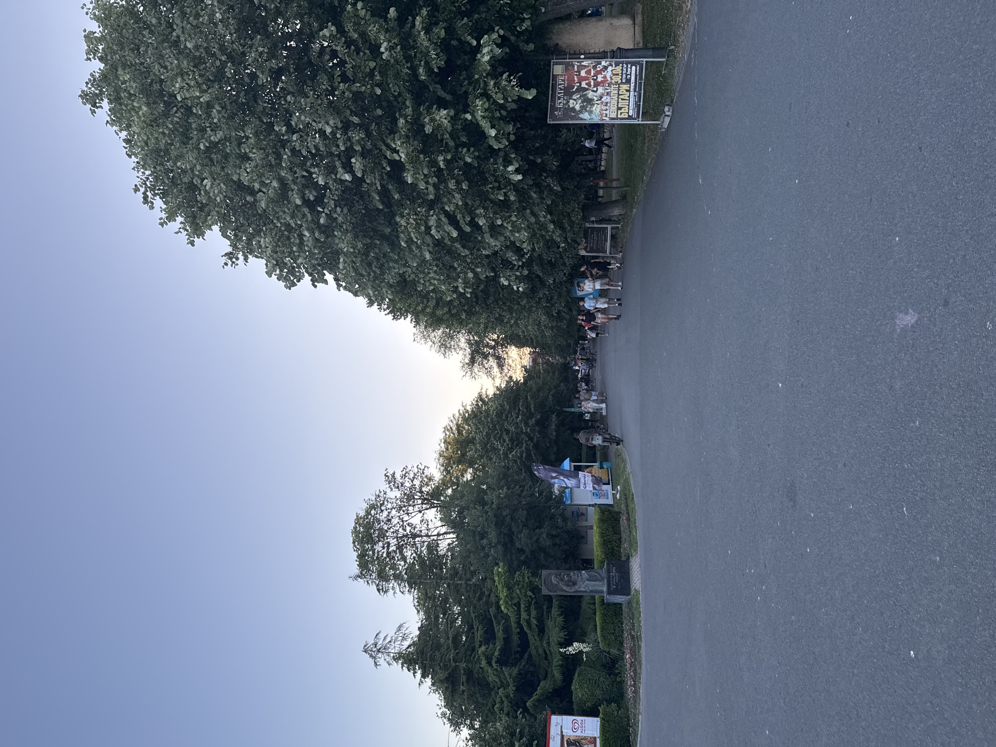 The image depicts an outdoor scene in a park-like environment. In the foreground, a paved pathway stretches into the distance, bordered by lush green trees and manicured bushes. Several individuals are walking along the pathway, and one person is riding a bicycle.
To the left, a statue is visible, and further back, there are booths or kiosks with signage, one of which advertises "ALGOIDA". To the right, a large billboard displays text and images, appearing to be an advertisement for an event or performance. The sky above is clear and a pale blue, suggesting it is either early morning or late afternoon.
The overall atmosphere is one of a public gathering space, likely a park or promenade in the city of Burgas, Bulgaria, given the presence of Bulgarian text on the billboard.