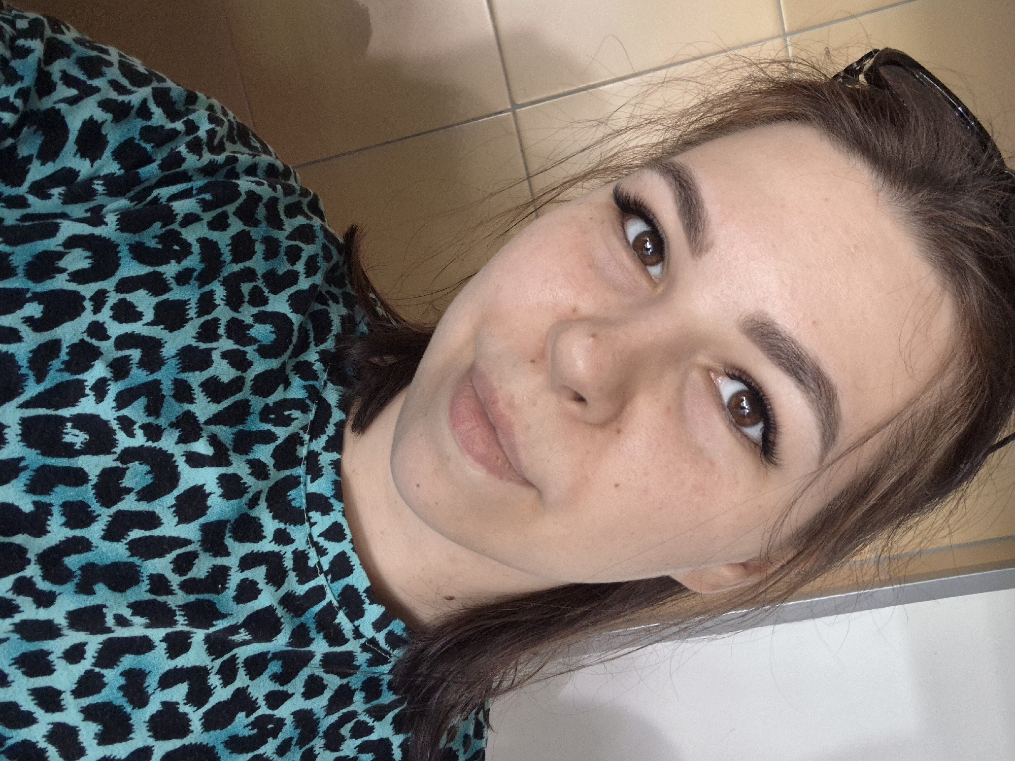 The image shows a selfie of a young woman with brown hair and brown eyes. She is wearing a blue and black leopard print shirt and has sunglasses perched on her head. She is indoors. There is no evidence to suggest the location is Tobolsk, Russia.