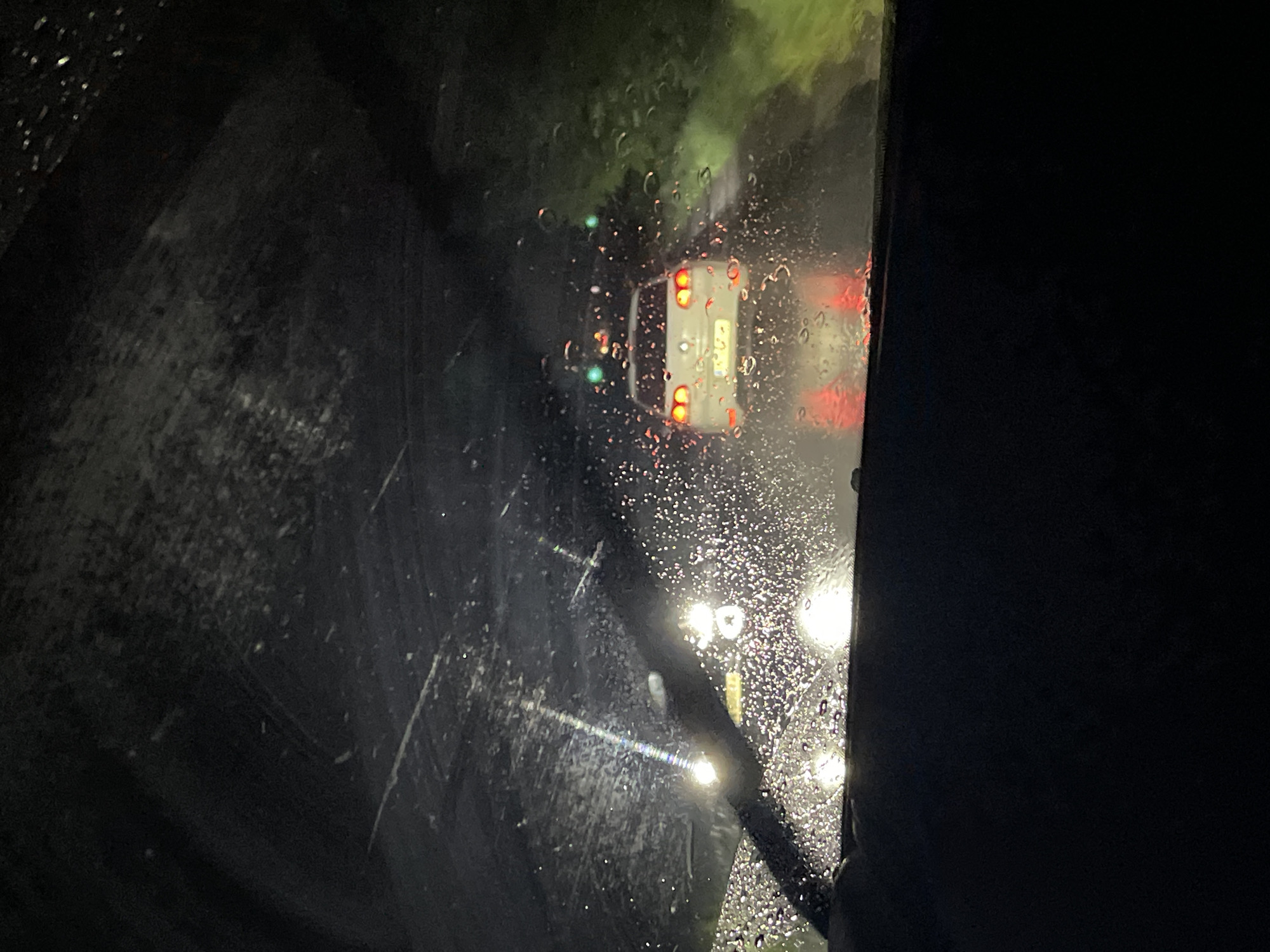 A nighttime view from inside a car during heavy rain. The windshield is covered in raindrops, with a wiper blade visible. Through the blurry windshield, the taillights of a white vehicle are seen ahead, along with streetlights. The location is likely a street in Batanovtsi, Bulgaria, though specific location details are not discernable from the image.
