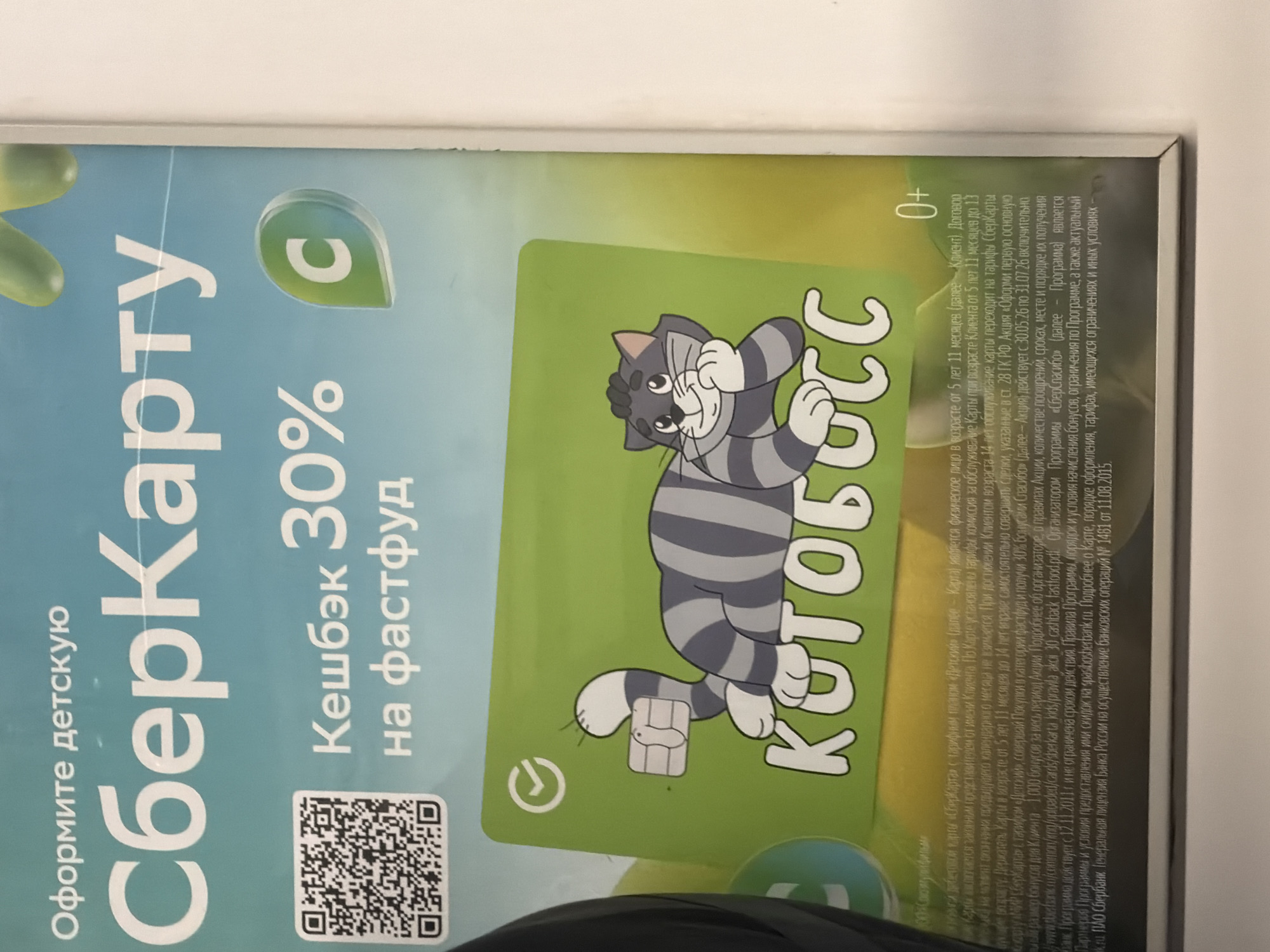 The image is a close-up photograph of a promotional poster for a Sberbank children's card. The poster features a cartoon cat on a bright green credit card with the text "КОТОБОСС" written on it. Above the card, there is text that reads "Оформите детскую СберКарту" (Apply for a SberKart children's card). To the left of this text, a QR code is visible. Further to the right, the text "Кешбэк 30% на фастфуд" (30% cashback on fast food) is displayed, along with a green circular logo containing the letter "C". The bottom of the poster contains smaller print with promotional details. The overall background is a gradient of light blue and yellow. The image appears to be taken in a well-lit indoor environment, possibly a retail space or an advertising display.