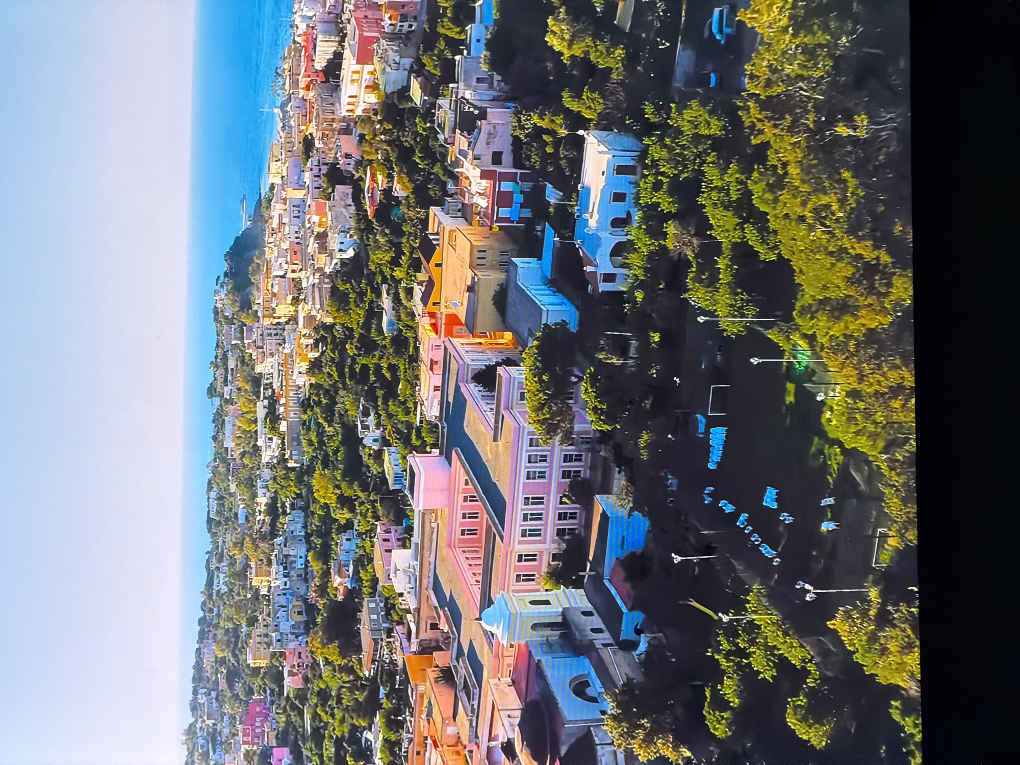 This aerial view captures a coastal town with a dense arrangement of buildings and lush green trees. The architecture features a variety of pastel colors, including pink, yellow, and white, with some structures exhibiting more vibrant hues like red. Several buildings appear to be residential, with multiple stories and visible windows. A prominent pink building with a symmetrical facade and numerous windows is centrally located in the foreground, accompanied by a white bell tower. Beyond these structures, the town stretches towards the sea, with the clear blue water meeting the horizon under a bright sky. The landscape is characterized by the pervasive greenery of trees that intersperse the urban development, suggesting a Mediterranean or similar climate. In the lower portion of the image, there is a sports field or open area with some objects and figures that are difficult to discern clearly due to the distance and resolution. The image does not provide any information suggesting the location is Chirchik, Uzbekistan.