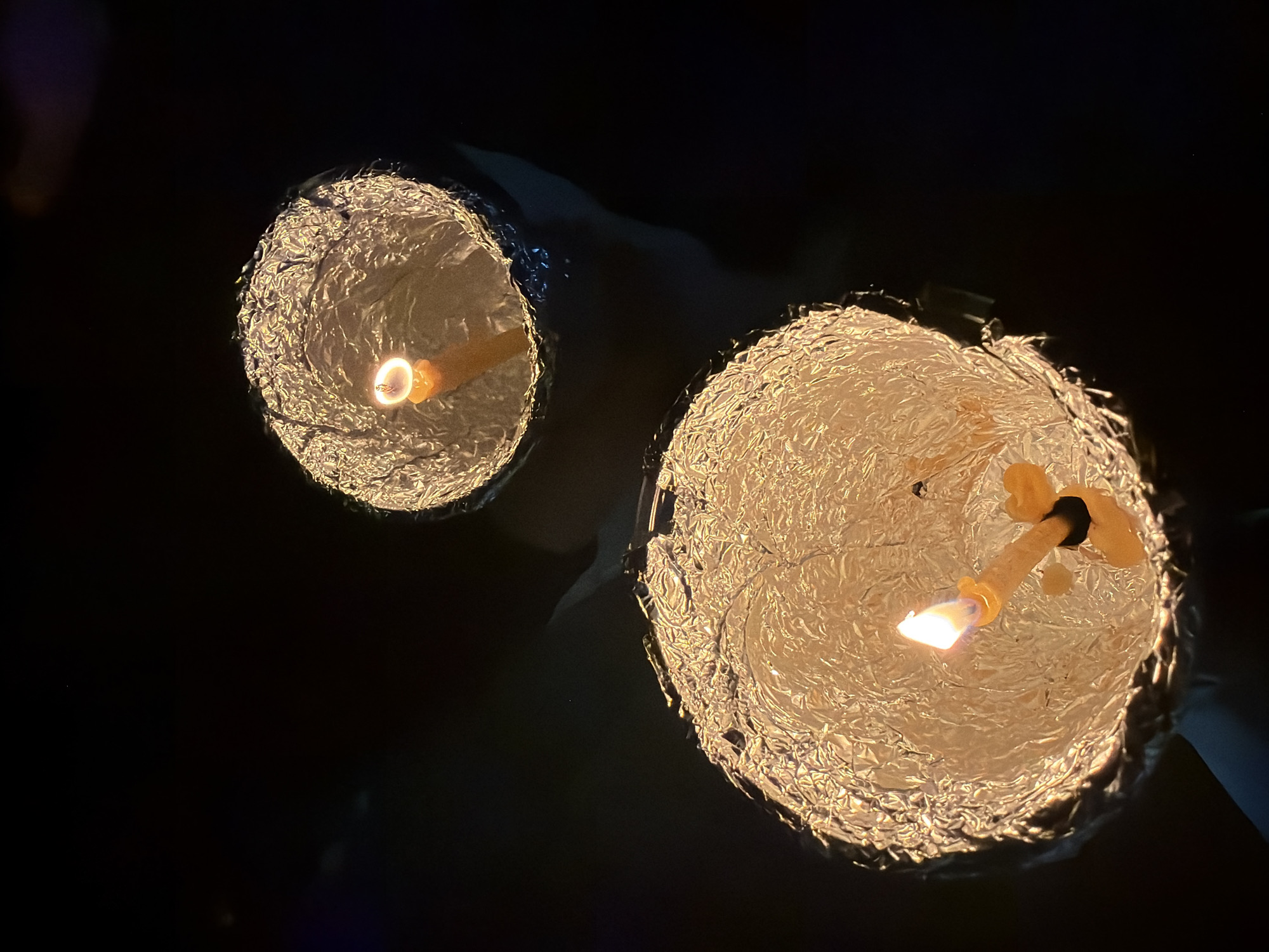 Two makeshift candle lanterns are illuminated in the dark. The lanterns are constructed from tin foil, appearing to have been molded into bowls or cups to contain the candles. The candle in the right lantern is larger than the left. There are hands or arms visible on the right of the image. The environment is predominantly dark. The location is unknown.