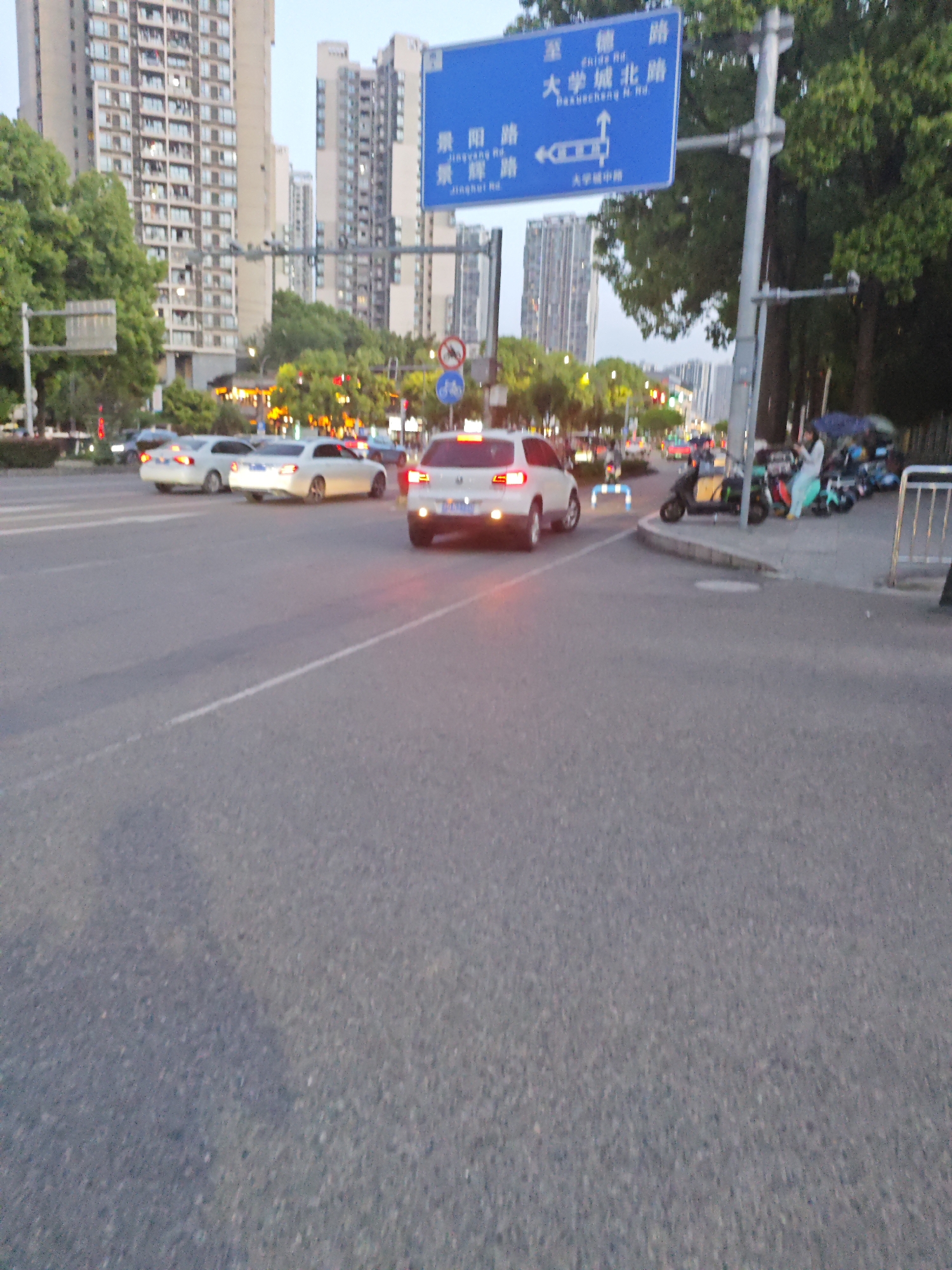 The image depicts a street scene in the city of Chongqing High-tech Zone, China. The foreground is dominated by an asphalt road. In the mid-ground, there are several cars and motorbikes present. A white car is positioned near the center of the frame, with its brake lights illuminated. A street sign is prominently displayed, indicating directions to "Zhuyin Road", "Daxuecheng N. Road" and "Jingyang Road". Buildings of considerable height, likely residential apartments, are visible in the background, and the sky above is overcast. A few trees are scattered throughout the scene. A group of people can be seen standing next to a row of parked motorbikes.