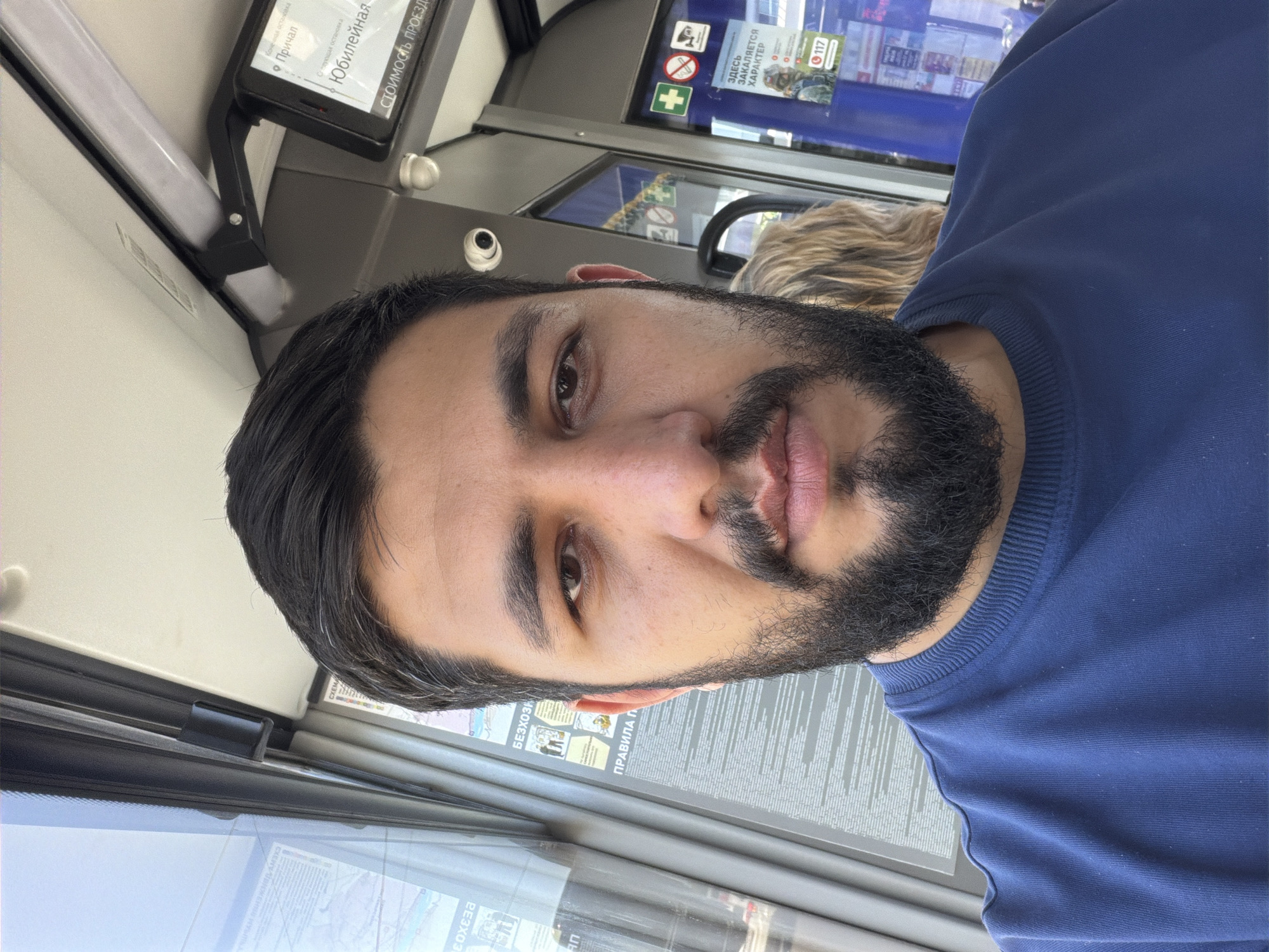 A young to middle-aged man with dark hair, a full beard, and a neutral expression is captured in a selfie-style photo, appearing to be inside a public transport vehicle. He is wearing a plain dark blue crew-neck sweatshirt.
The setting is the interior of what looks like a bus or tram, characterized by large windows and various mounted displays. Above and behind the man, an overhead digital screen displays information in Cyrillic script, including "Конечная остановка" (Final stop), "Причал" (Piers/Berth), and "Юбилейная" (Yubileynaya), which are likely names of stops. The phrase "СТОИМОСТЬ ПРОЕЗД." (Cost of travel/fare) is also visible on this display.
Further in the background, another sign features icons for security cameras, a no-smoking symbol, and a first-aid cross. Text on this sign includes "ЗДЕСЬ ЗАКАЛЯЕТСЯ ХАРАКТЕР" (Character is forged here) and the number "117". Below, through the window, reflections are visible, and more Cyrillic text can be seen on various notices, such as "БЕЗХОЗН" (part of 'unattended items') and "ПРАВИЛА П" (part of 'rules of conduct'). A bright fluorescent light fixture is installed overhead.
The presence of extensive Cyrillic text strongly indicates a location in Russia, consistent with the given context of Krasnoyarsk. The lighting suggests it is daytime. The man's expression is calm, and the activity appears to be a casual photo taken during a commute.