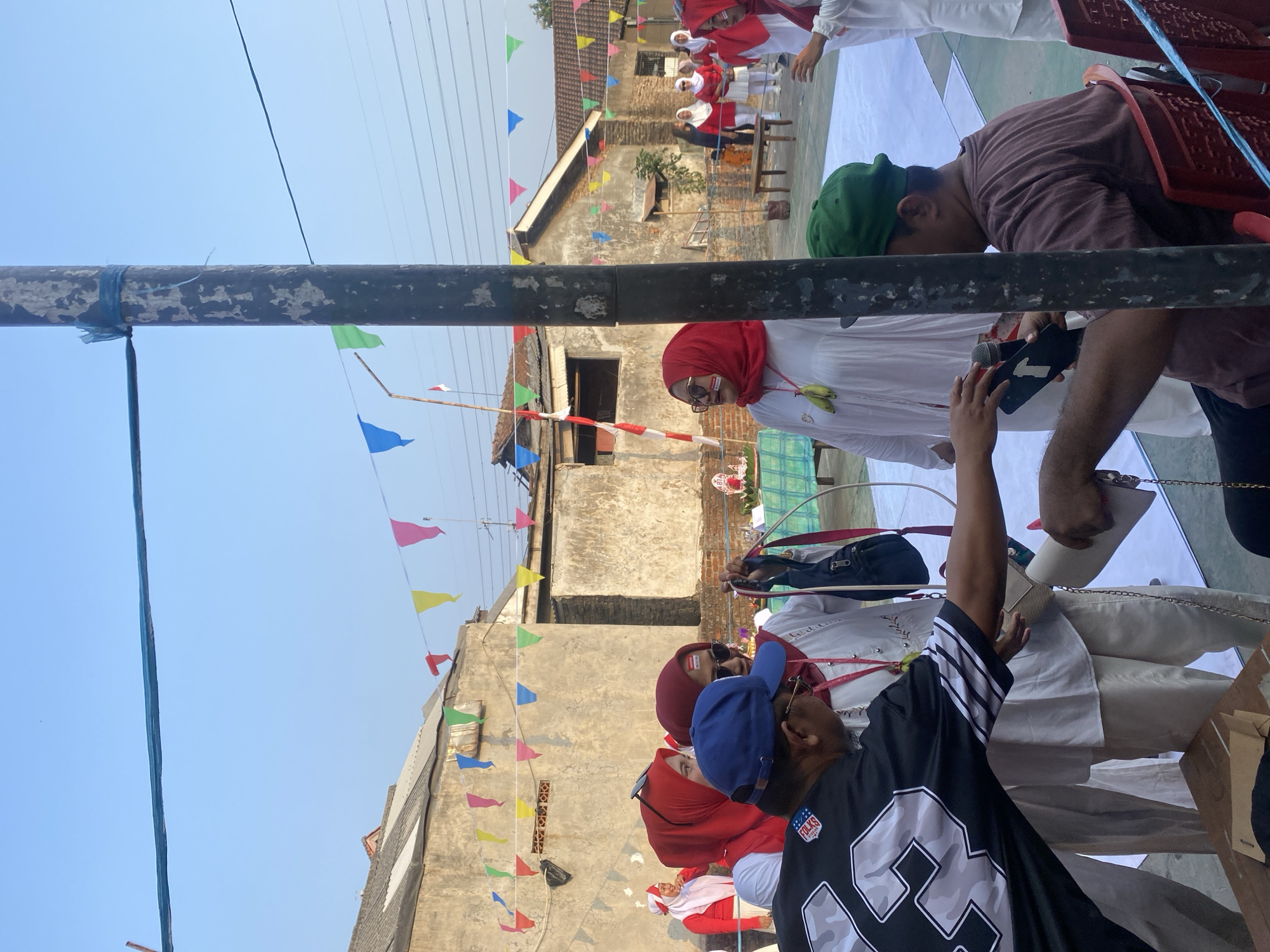 In the city of Cilegon, Indonesia, a group of people are gathered outdoors, possibly for a local celebration or event. The sky is clear and blue, and colorful triangular flags are strung across the scene, adding a festive atmosphere. In the foreground, a man wearing a black jersey with white and gray camouflage pattern and a blue baseball cap is interacting with a woman in a white outfit and a red headscarf. He appears to be handing her a phone or similar device. The woman is wearing sunglasses and has red and white markings on her face. To her right, another woman in a similar white outfit and red headscarf is also present, holding a dark blue bag.
In the background, a group of women in red headscarves and white attire are seen standing, possibly participating in the same event. There are also low-rise buildings with textured walls and tiled roofs, suggesting a residential or community area. A wooden table is visible, and a person wearing a green cap is seated in a red chair, looking away from the camera. Electrical wires crisscross the sky, and a utility pole with peeling paint stands prominently in the middle of the frame. The overall scene depicts a community gathering, likely in a warm, sunny climate, with cultural elements suggested by the attire and decorations.