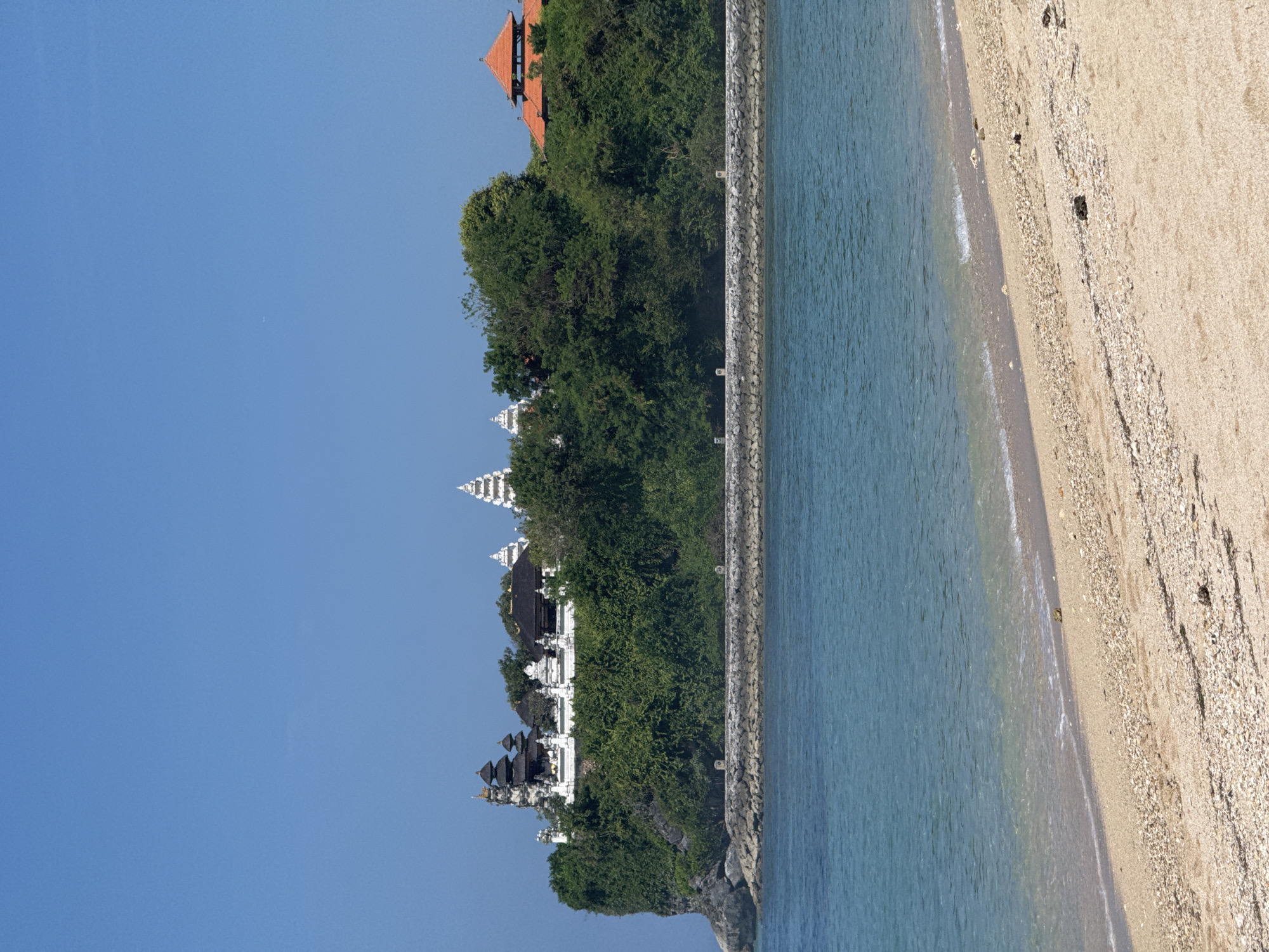 The image displays a serene coastal landscape under a clear blue sky. On the left side of the frame, a lush, verdant cliff or hill is covered in dense green trees. Perched atop this elevation are several traditional-style buildings. In the foreground on the cliff, there is a complex of white buildings featuring multiple-tiered, pagoda-like roofs, characteristic of temples or ceremonial structures, predominantly in white and dark grey. Further back on the hill, partially obscured by trees, stands a building with a distinctive red-tiled roof.
A long, grey, rocky barrier, resembling a sea wall or breakwater, extends horizontally across the middle of the image, separating the land from the water. To the right of this barrier, the calm, blue-green waters of the ocean gently lap at a sandy beach. The beach, composed of light-colored sand and scattered small pebbles or shells, curves slightly towards the foreground. Gentle waves are visible near the shoreline.
The scene suggests a bright, clear day, likely morning or early afternoon, with ample sunlight illuminating the landscape. There are no discernible human activities or visible text in the image. The overall impression is one of tranquility and natural beauty, highlighting traditional architecture in a picturesque coastal setting.