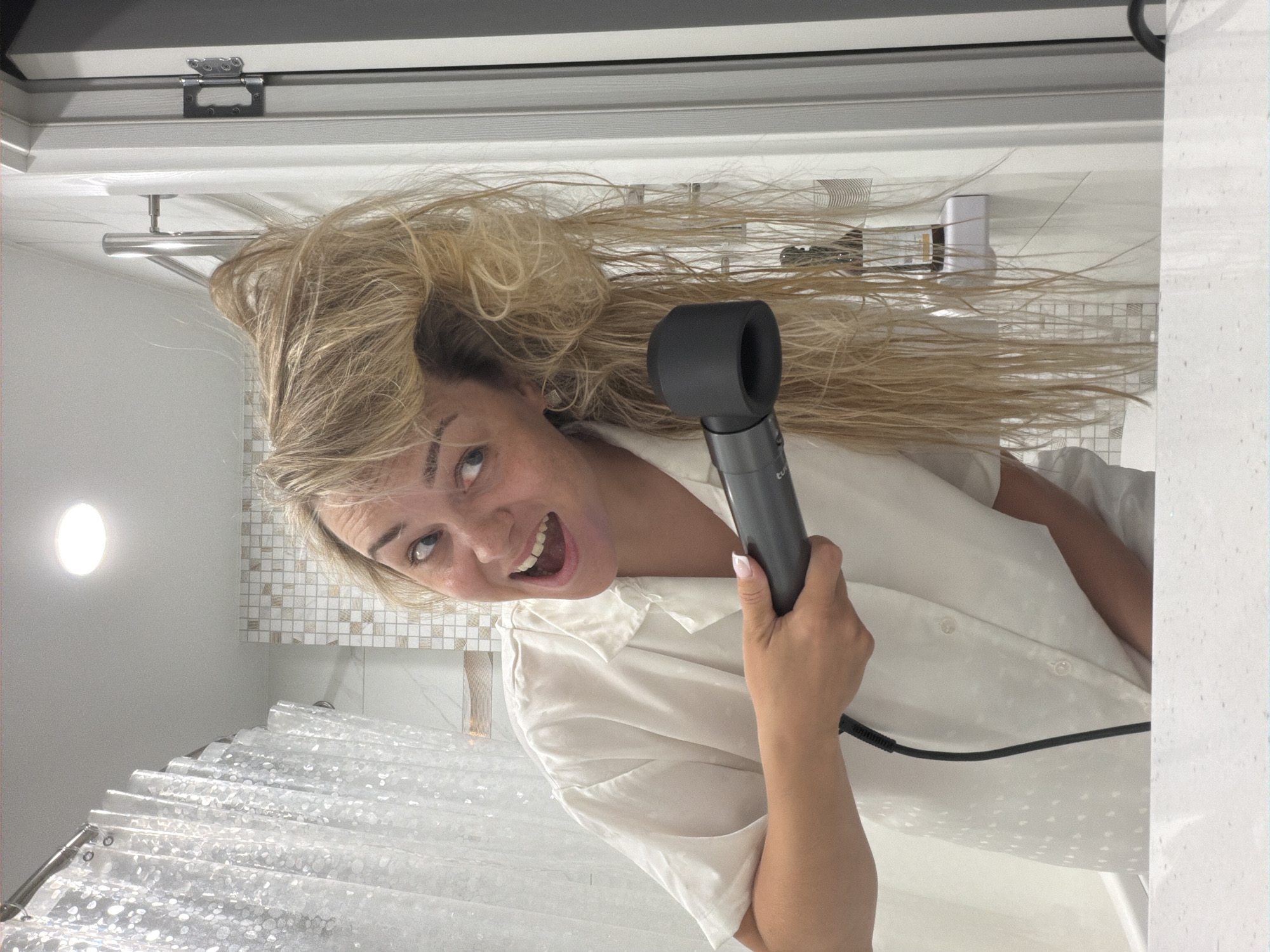A woman with blonde, shoulder-length hair is holding a Dyson hair dryer in a bathroom. She is wearing a white, button-down shirt and has a wide, open-mouthed smile. The bathroom has a white shower curtain, white and brown tile, and white walls and cabinets. The exact location within Votkinsk, Russia is indeterminable.