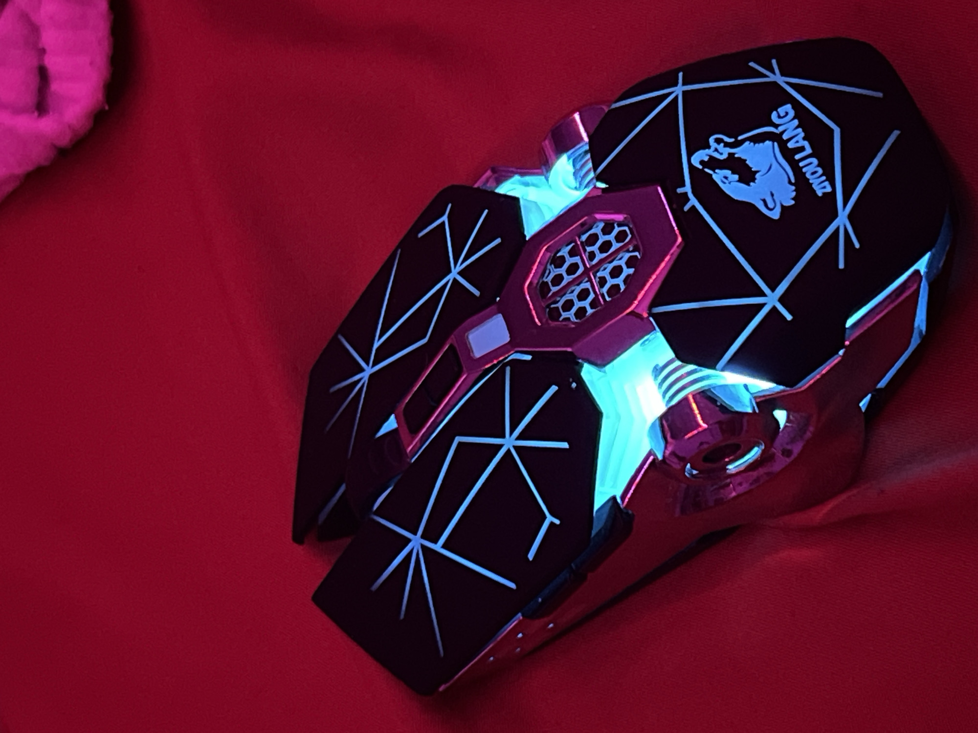 This image, reportedly captured in the city of Debelt, Bulgaria, displays a black gaming mouse with vibrant cyan LED illumination resting on a red fabric surface. The mouse features a futuristic design with geometric light-blue lines etched into its dark top panels. A central section of the mouse exhibits a honeycomb-patterned grill glowing with cyan light, flanked by metallic pink-hued accents. The right rear panel of the mouse prominently displays a glowing cyan wolf head logo above the text "ZIYOU LANG." Bright cyan light also emanates from various gaps and contours along the mouse's body. The background is a uniform red, soft-textured fabric, with a blurry, out-of-focus pink, fuzzy object partially visible in the top right corner. No specific architectural or environmental features identifying Debelt, Bulgaria, are discernible in the scene.