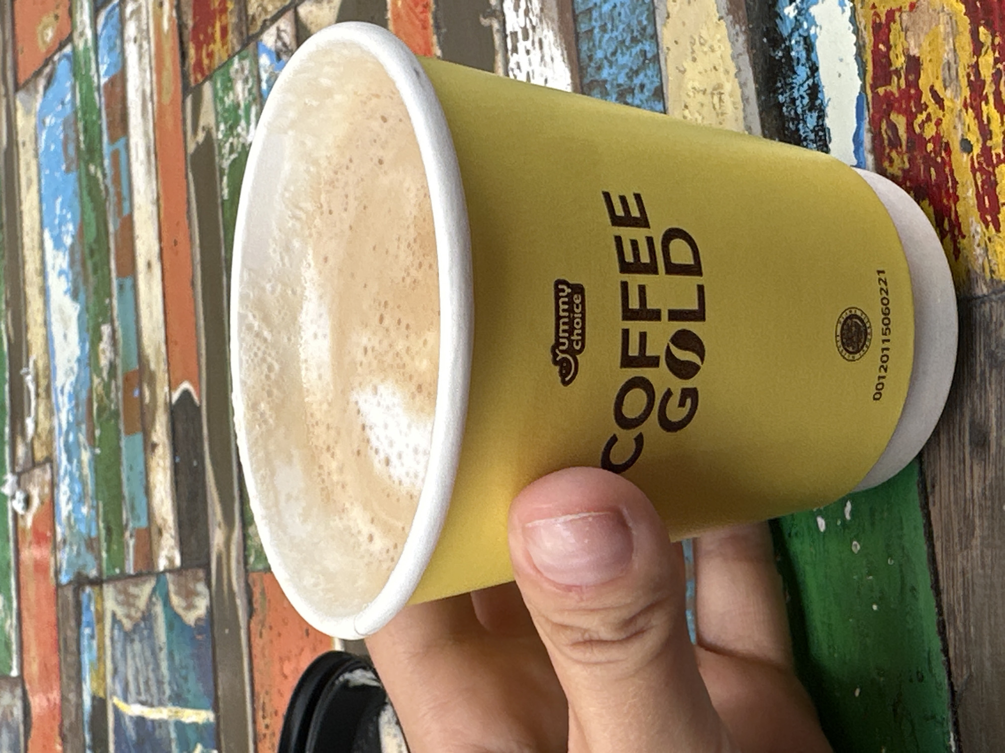 A close-up shot shows a hand holding a yellow disposable coffee cup filled with coffee, positioned on a colorful wooden surface. The cup has "Yummy choice" and "COFFEE GOLD" printed on it. The coffee surface is frothy. A black lid is partially visible on the left. The wooden surface features planks of various colors, including red, blue, green, and orange, arranged horizontally. The background is blurred. The scene appears to be outdoors or in a casual setting with rustic decor. There is no discernible city or location information available.