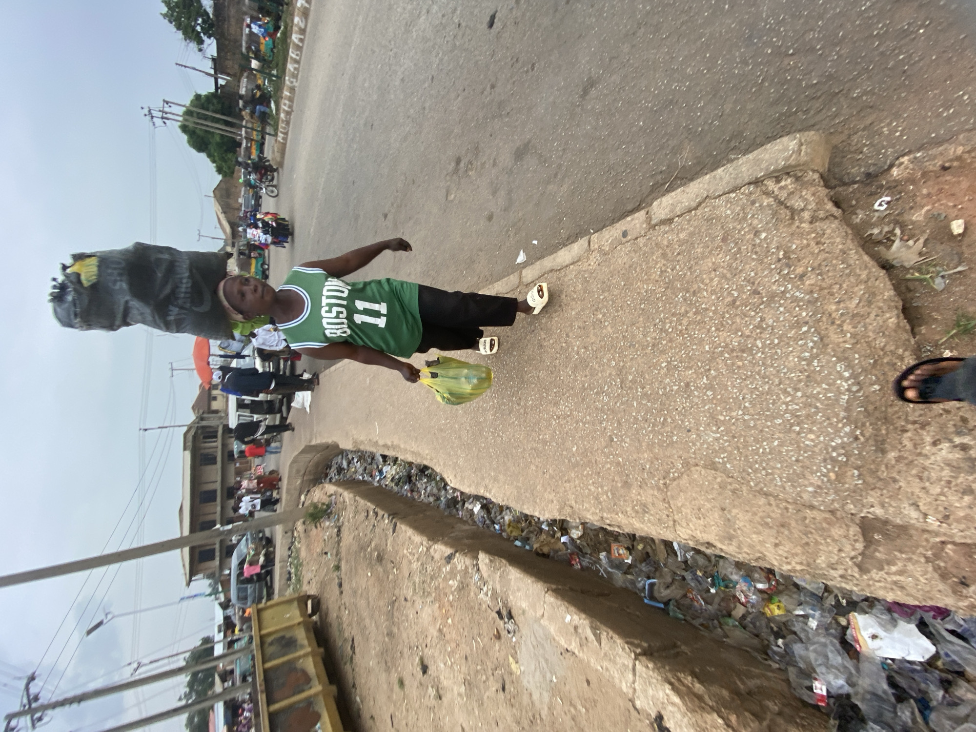 This daytime image captures a street scene in Bukuru, Nigeria. The primary subject is a person standing on a concrete sidewalk, wearing a green basketball jersey with "BOSTON" printed horizontally and "11" vertically on the front. They are also wearing black shorts or pants and white sandals, which show a faint "adidas" logo. The person holds a translucent yellow plastic bag in their right hand.
To the left of the person, on the sidewalk, stands a large, dark, upright object, possibly a statue or a heavily wrapped item, with the text "Wey AncheW" visible on its side. The sidewalk runs alongside a paved road, which features painted white text that appears to be "HUCHIAGASAZT" or similar.
A notable detail is the open ditch or gutter that separates the sidewalk from a lower earthen area, completely filled with an extensive collection of plastic waste and other debris. In the background, the street is busy with people and vehicles, including motorcycles and tricycles. Multi-story buildings line the road, and power lines crisscross the sky. A large, yellow, open-top container, resembling a dumpster, is partially visible in the lower left, and an orange umbrella can be seen further down the street. The sky appears overcast or hazy.