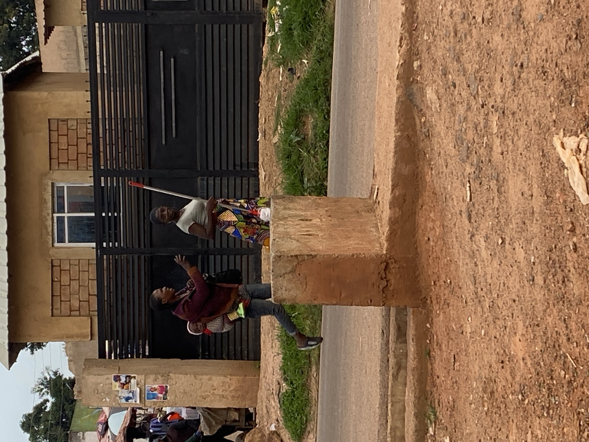 In the city of Bukuru, Nigeria, a woman is captured walking with a baby strapped to her back. She is wearing a maroon jacket over a white and striped top, dark denim jeans, and black sandals. The child on her back is wearing a striped garment. Another woman, to her right, is wearing a light green t-shirt and a colorful patterned skirt, holding a red-handled broom. They are walking on a dusty, reddish-brown street next to a raised concrete structure and a black metal gate. Behind the gate, a building with a light brown facade and a corrugated metal roof is visible. The environment appears to be a public area, possibly near a marketplace, given the presence of stalls and people in the background to the left. The overall lighting suggests it is daytime, possibly overcast.