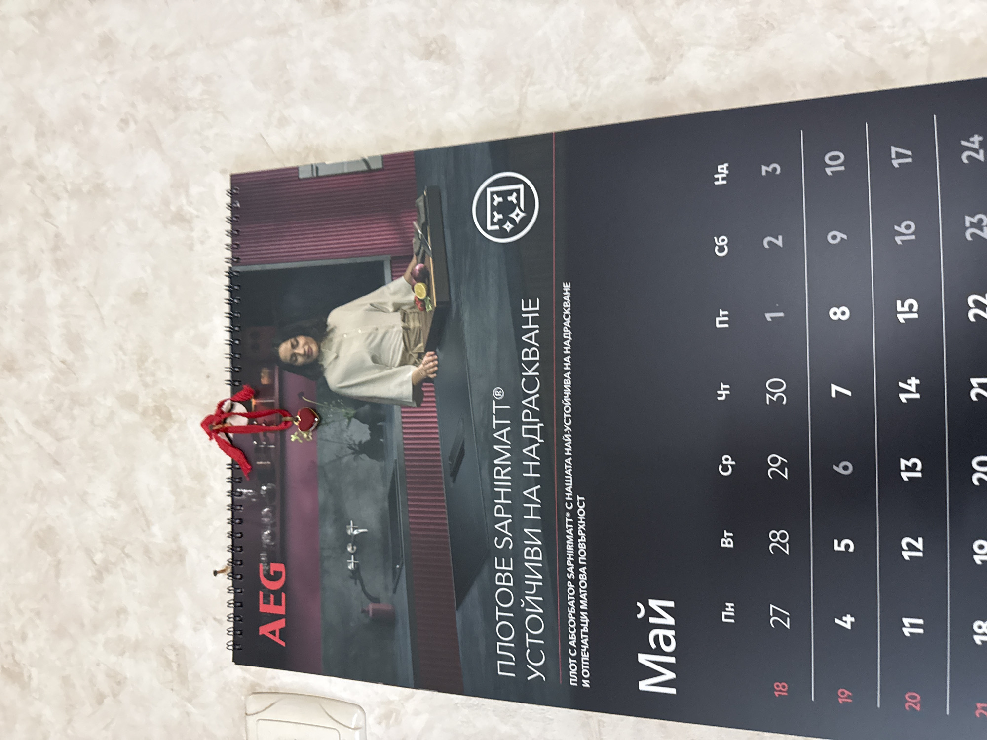 Here is a description of the image:
The image shows a spiral-bound calendar lying on a textured surface. The calendar is open to the month of May. The brand "AEG" is prominently displayed on the left side of the calendar. An image shows a woman beside what is presumably a "SaphirMatt" countertop. The text on the calendar, written in Bulgarian, advertises "SaphirMatt" countertops as being scratch-resistant. The calendar grid shows the dates for May, starting with the 18th. The calendar appears to be near the city of Lovech, Bulgaria, based on the Bulgarian text used.