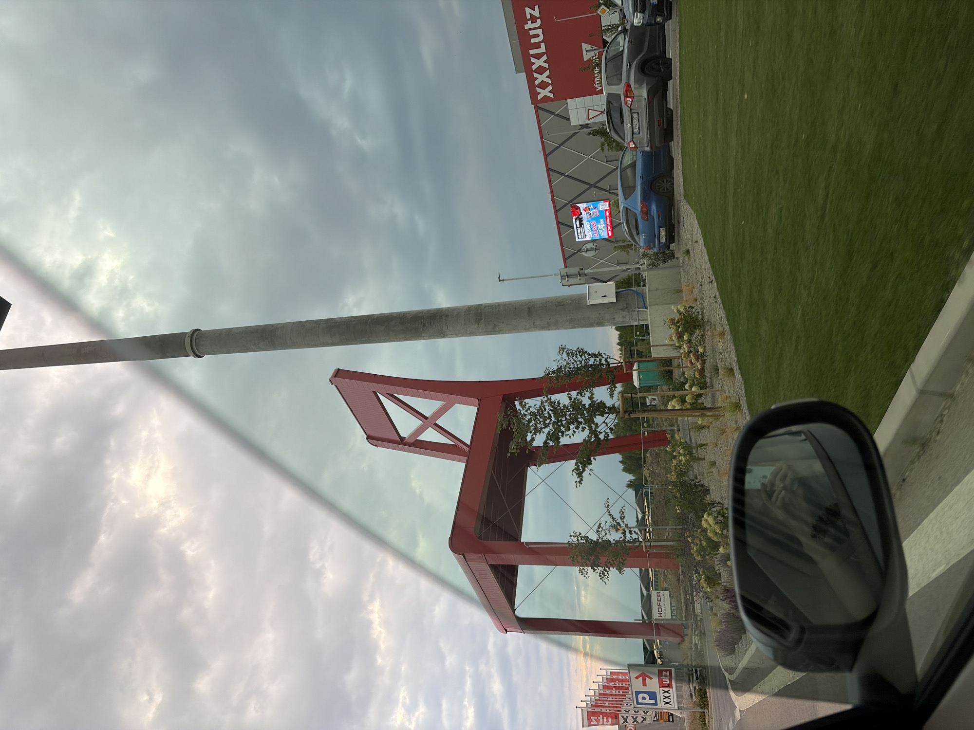 In Nitra, Slovakia, a large, vibrant red chair sculpture stands prominently against a cloudy sky. The sculpture is situated in front of a modern building complex with a red facade bearing the XXXLutz logo. To the left of the sculpture, several red and white flags are visible, along with a parking sign indicating "P" and "XXX Lutz". Cars, including a blue Mazda and a grey Suzuki, are parked in front of the XXXLutz building. The scene is captured from the perspective of a moving vehicle, with the reflection of the car's interior, including a person's hands holding a phone, visible in the side mirror. The foreground shows a neatly mowed green lawn and a concrete barrier.