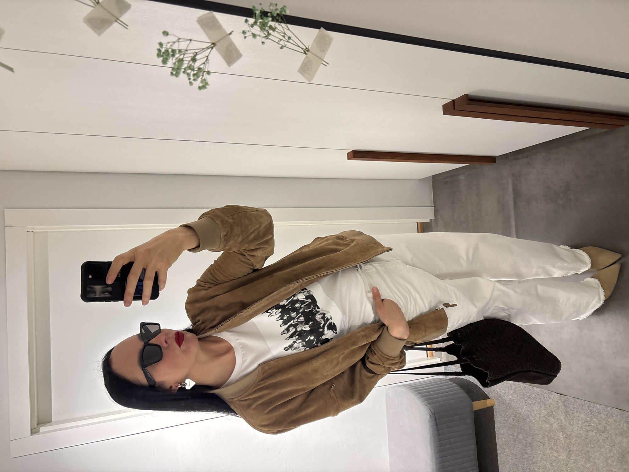 A woman is taking a selfie in what appears to be a fitting room. She is wearing a brown suede bomber jacket, a white graphic t-shirt, and white trousers. She has black hair, is wearing sunglasses, and has a red lip. She's holding a black smartphone in her left hand, and the phone case features an image. To her right is a gray padded stool and a dark-colored handbag. The room has white walls and a gray floor. The ceiling is white with black trim and has faux greenery attached with tape. Wooden ledges are mounted on the wall above the fitting room door. The city of Chaikovsky, Russia, is not directly identifiable from the image.