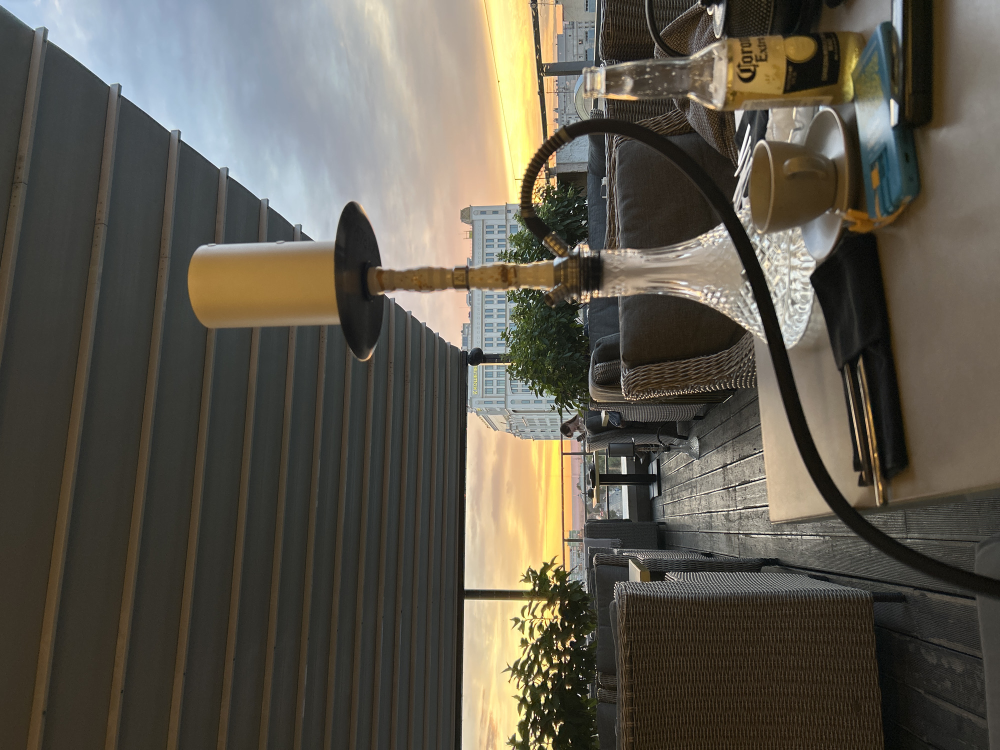 This is a vertical, eye-level, medium shot taken from a rooftop terrace at dusk, overlooking a city skyline and a sunset. The setting appears to be a cafe or restaurant with outdoor seating, furnished with wicker chairs and tables. A hookah is prominently featured in the foreground, with its glass base and hose visible. On a table to the right, there is a Corona Extra beer bottle, a teacup and saucer, and a cell phone.
The sky is a gradient of warm colors, transitioning from yellow and orange near the horizon to pale blue and grey higher up, indicating the time of day is sunset. The cityscape in the background consists of several buildings, some of which are illuminated, suggesting it is an urban environment. The presence of outdoor furniture and the relaxed atmosphere, coupled with the sunset, evoke a sense of leisure and enjoyment.
A person is visible in the background, seated at a table, further suggesting this is a social or recreational space. The weather appears clear, with no indication of rain or clouds other than those naturally present in the sky during sunset. The visible text on the beer bottle reads "Corona Extra". The overall mood is calm and serene, enhanced by the soft lighting of the setting sun. The location context is Novokuybyshevsk, Russia.