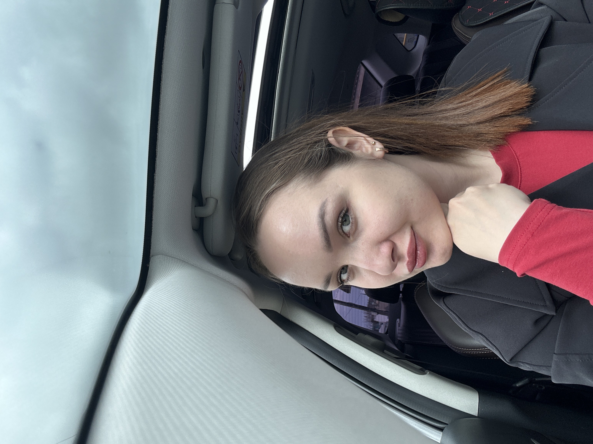 A person is inside a car, positioned near the sunroof. The person has light skin, light eyes, and shoulder-length brown hair. The person wears a red shirt and a black jacket. The interior of the car is primarily gray and black. The location is potentially Chaikovsky, Russia, but there is no visible information that confirms this location.