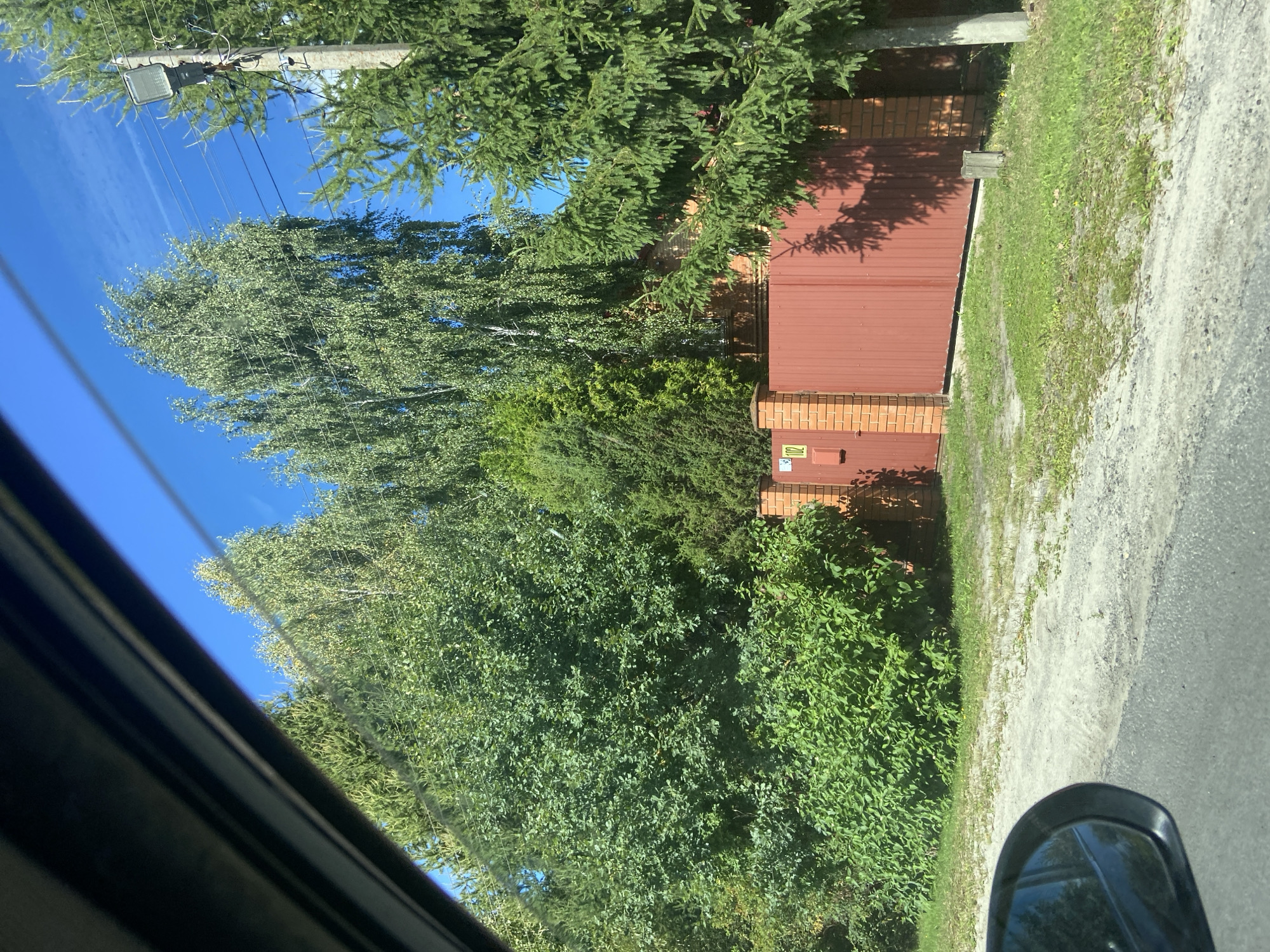 This image was taken from inside a vehicle looking out at a residential property. The foreground shows the car's interior, with a black frame along the top left and a side-view mirror on the bottom left. The exterior view features a large, red metal gate with brick posts and a small sign with the number "102" on the left side. Behind the gate, dense green foliage and trees are visible, including a tall birch tree and a coniferous tree. The property appears to be situated in a rural or suburban area, with a dirt or gravel road in front of the gate and a paved road in the foreground. The sky is clear and blue. This image is located in Astashkovo, Russia.