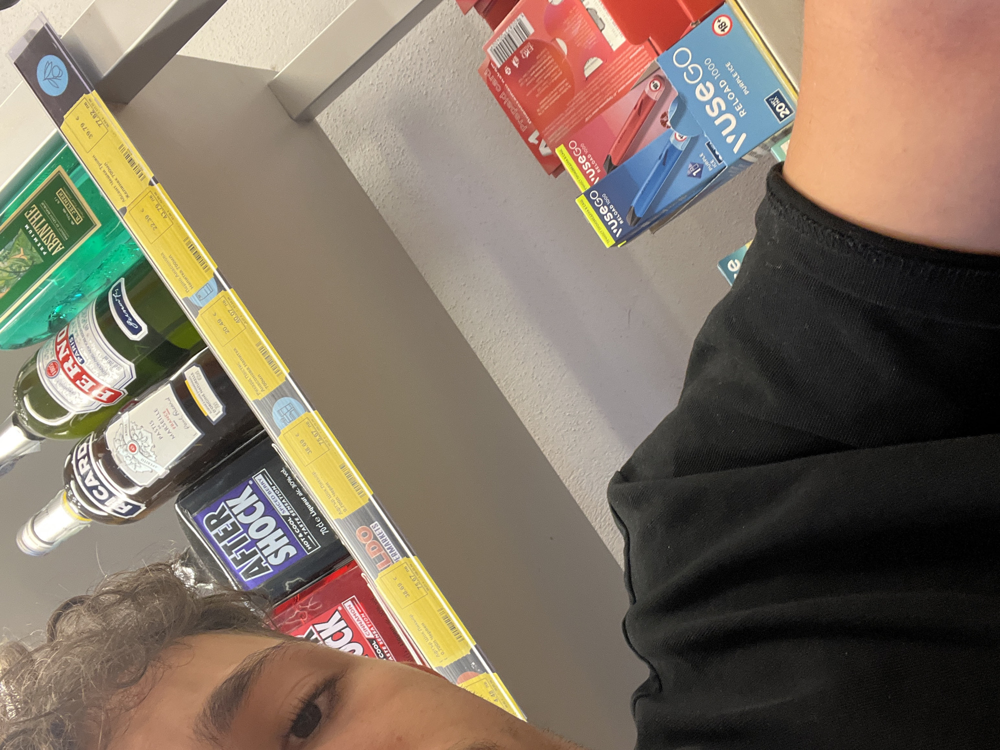 A close-up, slightly distorted selfie of a person's face and shoulder is captured in a brightly lit retail environment, likely a supermarket or liquor store in Varna, Bulgaria, given the Cyrillic text on the product labels. The person, with curly brown hair and visible eyebrows, is wearing a black t-shirt. The background features a shelf stocked with various alcoholic beverages, including bottles of Ricard Pastis Marseille, a green bottle labeled "PREMIUM ABSINTHE," and a black bottle of "AFTER SHOCK" liqueur. Price tags with Cyrillic and numerical denominations are attached to the shelf. Further down the shelf, several boxes of "vuse GO" vape products are visible, one blue and one red. The overall impression is one of casual shopping.