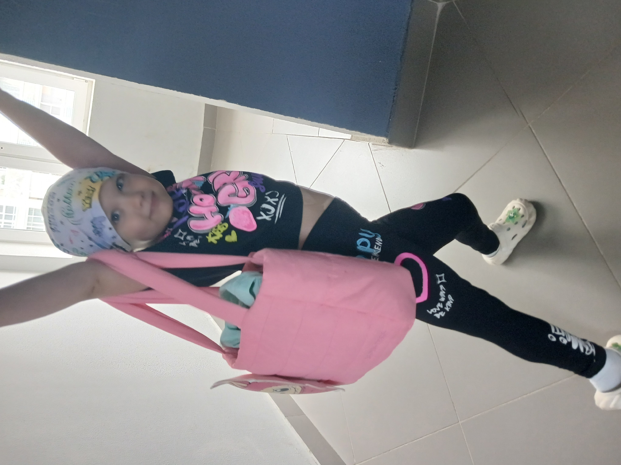 A young girl with a bandana on her head and a bright, colorful graphic t-shirt is lying on her back on a tiled floor. She is holding a pink bag with a light blue item inside. The girl is wearing black leggings with white text and graphics, and white Crocs with green charms. Her arms are outstretched, and she is looking up at the camera with a smile. The scene appears to be indoors, possibly in a hallway or common area, with a blue wall in the background and large windows visible on the left. The lighting suggests it is daytime. The overall impression is one of playfulness and joy. Visible text on the t-shirt appears to be "XOXO". Visible text on the leggings includes "LOVE WANTS BE KIND" and possibly other stylized writing. The setting suggests a casual, everyday environment.