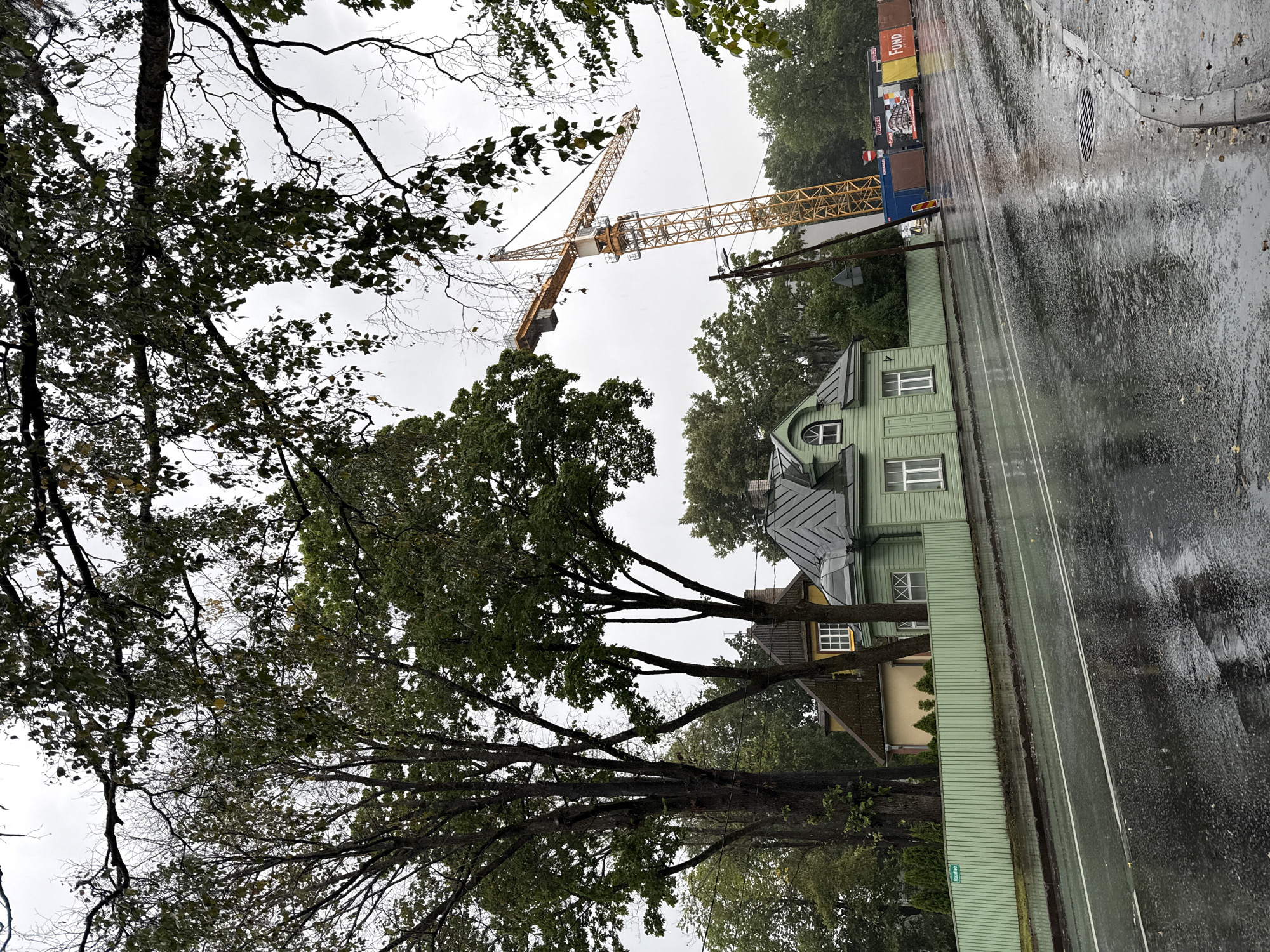 The image shows a street in Tallinn, Estonia, on a rainy day. The foreground is dominated by a wet asphalt road with reflections of the surroundings. A green wooden fence runs along the side of the road, separating it from a property with a light green house and a yellow house behind it. In the background, a large yellow construction crane is visible, along with shipping containers and construction site signage. Trees with green foliage frame the top of the image. The sky is overcast.